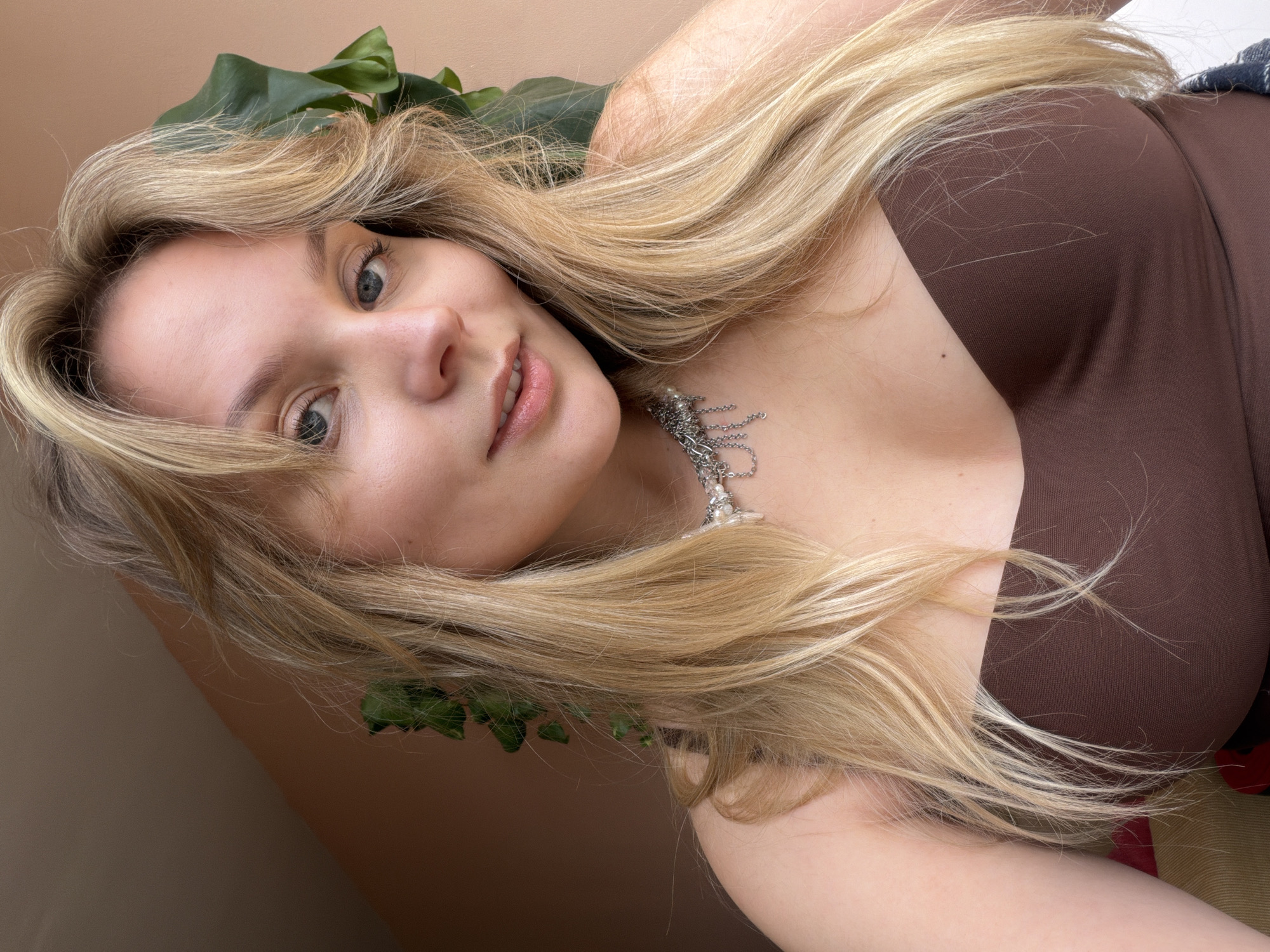 The image displays a close-up, frontal portrait of a young adult female. She possesses fair skin, light blue eyes, and long, wavy light blonde hair with face-framing layers and bangs. Her lips are slightly parted. She is wearing a brown strapless top and a multi-strand silver-toned necklace adorned with white pearls and metallic letter-shaped charms. A large green leaf, consistent with a houseplant, is visible behind her right shoulder and head. The background features a light peach-colored wall, with a distinct darker brown diagonal section present in the upper left portion of the frame. The subject is looking directly into the camera. This scene is an indoor setting located in Tel-Aviv, Israel.