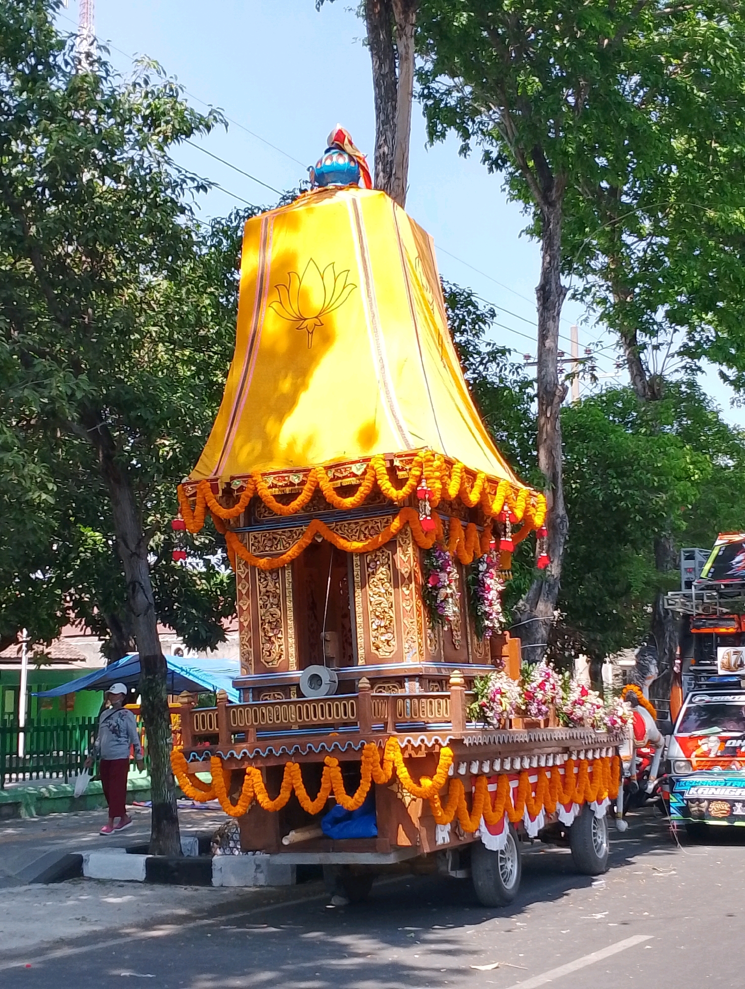 In Probolinggo, Indonesia, a brightly decorated chariot is displayed during a procession. The chariot features a tall, yellow, conical canopy adorned with a lotus design and intricate gold detailing. Orange flower garlands and red fabric hang from the canopy's edges. The chariot itself is made of dark wood with ornate carvings and is decorated with more orange garlands and colorful floral arrangements. The chariot is mounted on a flatbed truck with visible wheels and is part of a parade, as evidenced by another truck decorated with banners and lights visible to the right. A person in red pants and a grey shirt is walking near the chariot on the left side of the image. The scene is set outdoors, with lush green trees and a clear blue sky in the background, suggesting a sunny day.