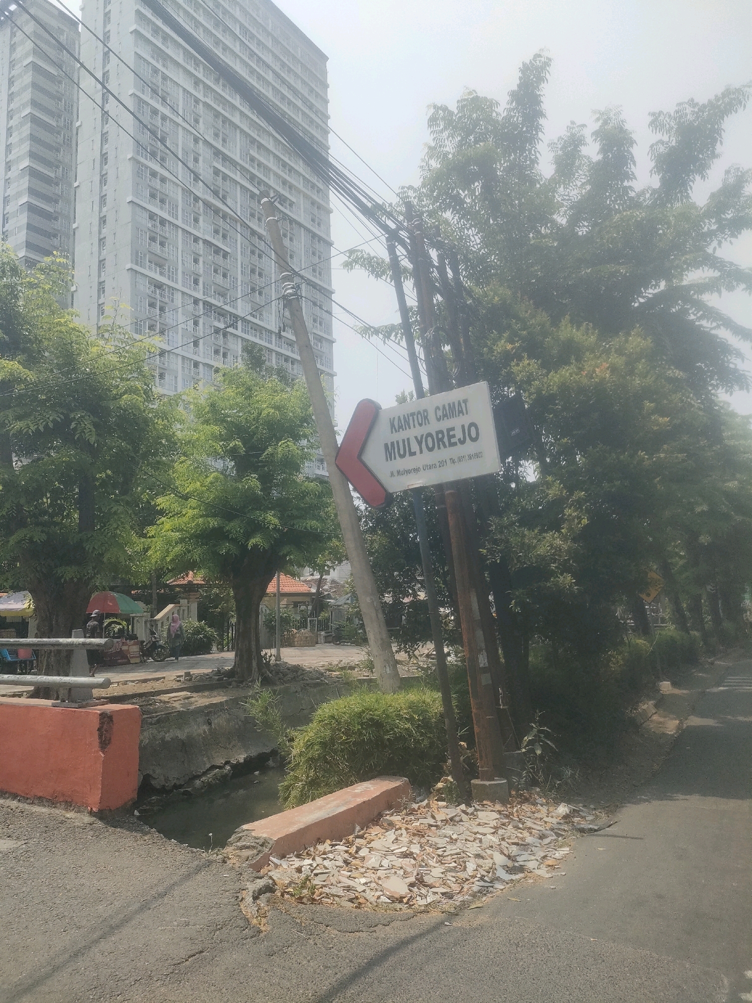 In Surabaya, Indonesia, on a hazy day, an outdoor scene unfolds. A tall, modern apartment building stands in the background, its numerous windows reflecting the muted sky. In the mid-ground, a road stretches into the distance, lined with lush green trees. A directional signpost, pointing left, reads "KANTOR CAMAT MULYOREJO" and provides an address "JL. Mulyorejo Utara 201. Tlp. (031) 5919002". The sign is mounted on a wooden pole, which is part of a larger assembly of utility poles and wires that crisscross the frame. A narrow, murky canal runs alongside the road, bordered by low concrete structures. The canal's edge is covered with debris, including broken tiles and scattered rubble. On the left side of the road, a red-painted concrete structure, possibly part of a bridge or embankment, juts out. Further down the road, several food stalls with colorful awnings are visible, along with a parked motorcycle. A person wearing a headscarf walks in the distance, away from the camera. The overall atmosphere is urban, with elements of residential development, public services, and commercial activity.