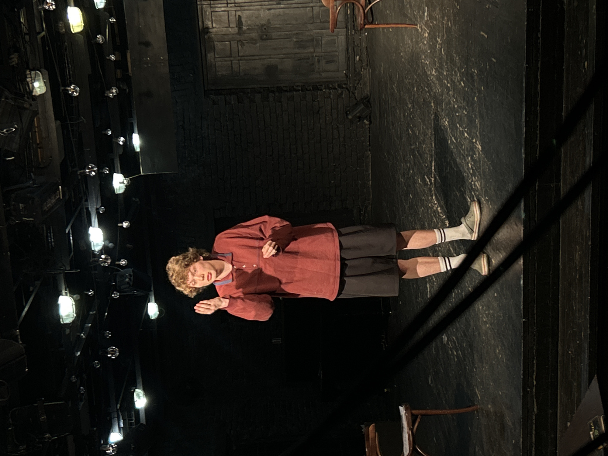 A person stands on a dimly lit stage, likely performing or rehearsing. The subject is a young individual with curly, light brown hair, wearing a reddish-brown long-sleeved polo shirt, dark shorts, and light-colored sneakers with white striped socks. They are captured mid-gesture, with their left hand raised, palm open, and their mouth slightly agape, suggesting they are speaking or singing.
The setting appears to be a theater or performance venue. The backdrop consists of a dark, textured wall, possibly painted to resemble brickwork at the bottom and wooden panels or a flat at the top. Numerous stage lights, both practical fixtures and small reflective spheres, hang overhead, casting light and shadows across the scene. The stage floor is dark and somewhat scuffed, reflecting some of the light. Two simple wooden chairs are partially visible on the stage; one is in the upper right corner, and another is in the lower right. The overall atmosphere is dark, with the focus illuminated on the performer, typical of a theatrical production. No visible text is present.