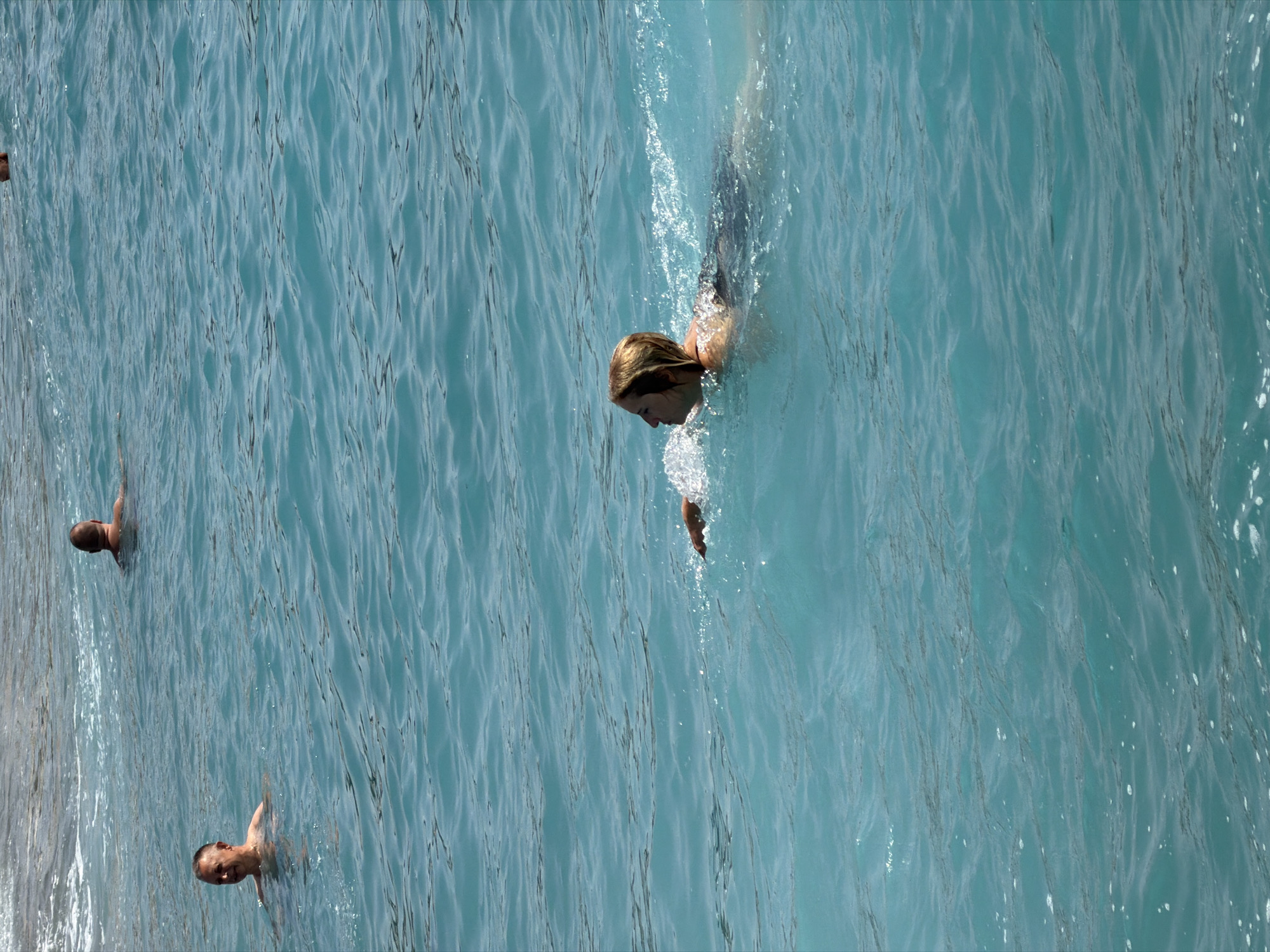 In the turquoise waters off Prijevor, Montenegro, four individuals are engaged in swimming. In the foreground, a woman with blonde hair is swimming towards the left of the frame, her body partially submerged and creating a noticeable wake. To her left and further back, a man with short brown hair is also swimming, facing left. In the middle ground, another man, this one with a shaved head, is swimming away from the viewer. The background reveals a hint of a sandy shore with gentle waves lapping at the coastline. The water is clear and exhibits a vibrant blue-green hue, with sunlight glinting off its surface.