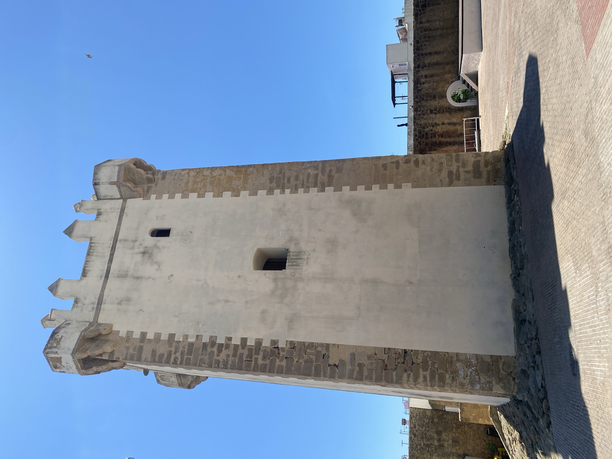The image captures a square, whitewashed tower with stone battlements and rectangular cutouts against a clear blue sky. The tower is located in the city of Conil de la Frontera, Spain, specifically in the Torre de Guzmán. The tower is made of stone and whitewashed plaster, with distinct crenellations along its upper edge. Two small, rectangular windows are visible on the main face of the tower. The base of the tower is surrounded by a cobblestone ground and some rubble. In the background, a low, dark stone wall stretches across the scene, topped with modern buildings and structures. A small, arched entrance with steps is visible in the wall. The lighting suggests it is daytime, with the sun casting a long shadow from the tower onto the foreground. No people are clearly visible.
