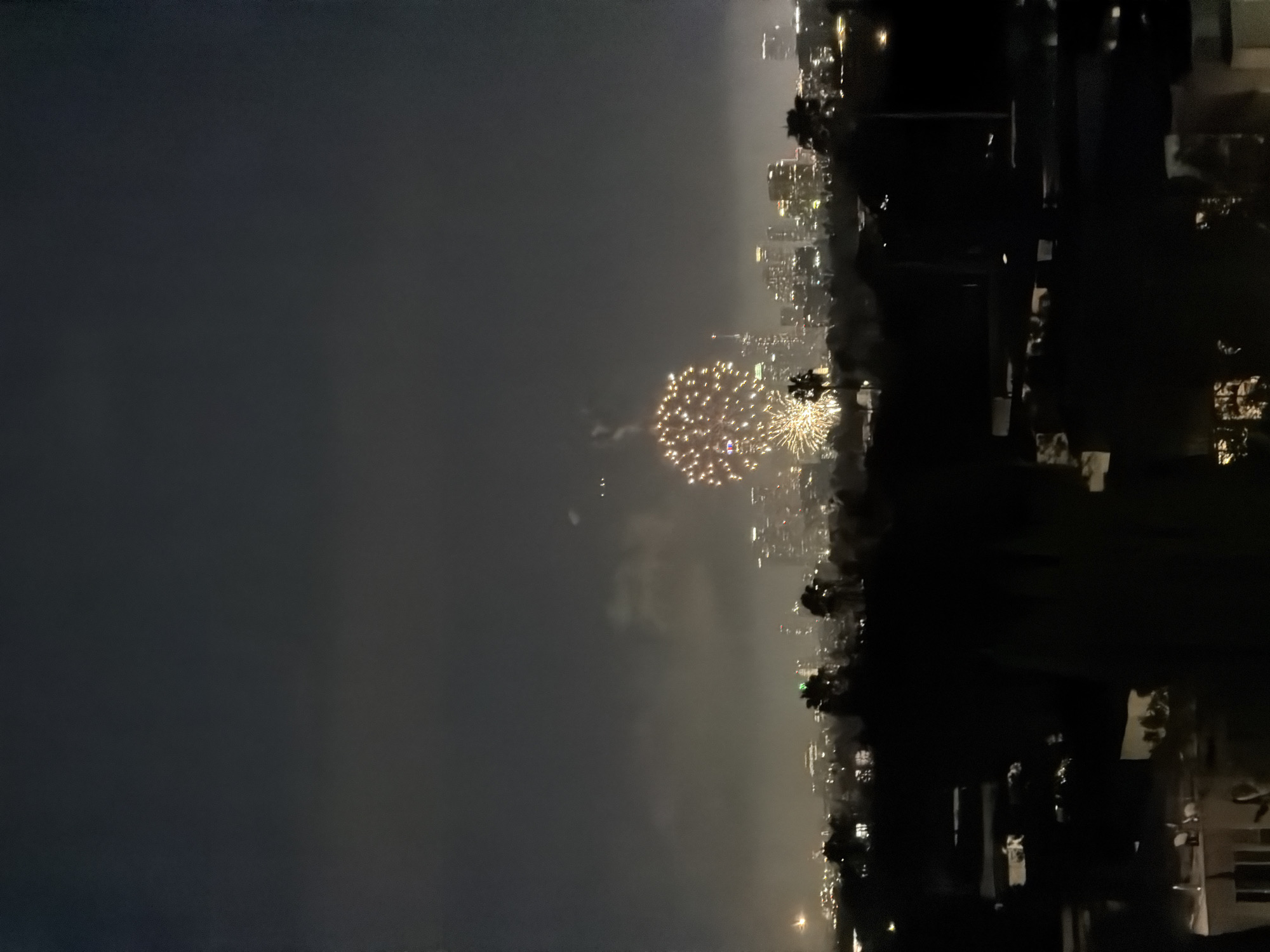 A nighttime cityscape view shows fireworks exploding over a city skyline. The foreground is filled with silhouetted trees and structures, possibly houses or buildings, obscuring the lower portion of the scene. In the mid-ground, a dense collection of buildings forms the city skyline, with lights visible from windows and streetlights. Towering over the skyline are several skyscrapers, their tops partially obscured by haze or smoke. The main subject of the image is a large burst of golden fireworks in the upper center, its light illuminating the sky and the surrounding smoke. Another smaller cluster of fireworks is visible to the left of the main display. The sky is dark and cloudy, with a slightly hazy atmosphere that softens the lights of the city. The location is the city of West Hollywood, United States of America.