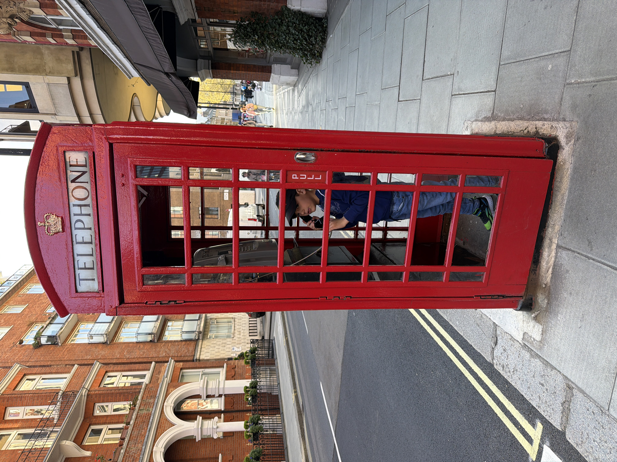 A slightly tilted, eye-level, daytime shot shows a child inside a red telephone booth, with the architecture and street scene of London, UK, visible in the background.
The classic red telephone booth stands on a gray stone sidewalk bordering a street marked with double yellow lines. A young boy inside the booth is engaged with the telephone handset. He wears a dark baseball cap, a blue long-sleeved shirt, jeans, and multicolored sneakers.
Behind the booth, across the street, is a row of brick buildings with multiple windows and ornate white detailing. Other buildings with awnings and various architectural styles are visible. The scene is set on a partly sunny day, with pedestrians visible further down the sidewalk in the background.