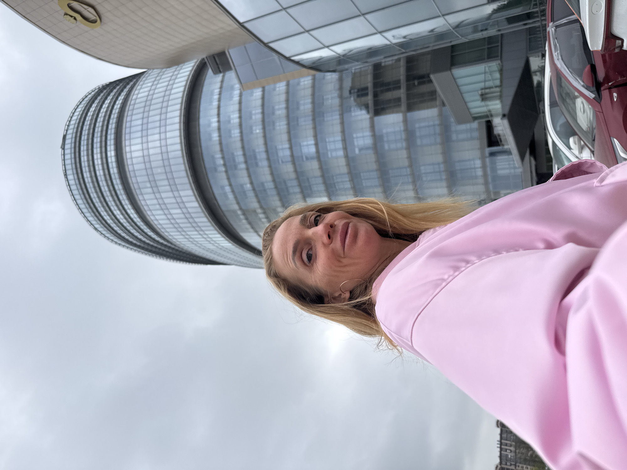 The image shows a woman in a pink jacket looking upwards at the Lakhta Center in Saint Petersburg, Russia. The Lakhta Center is a modern skyscraper with a curved, glass façade. Behind the woman, a maroon vehicle is partially visible. The sky is overcast.