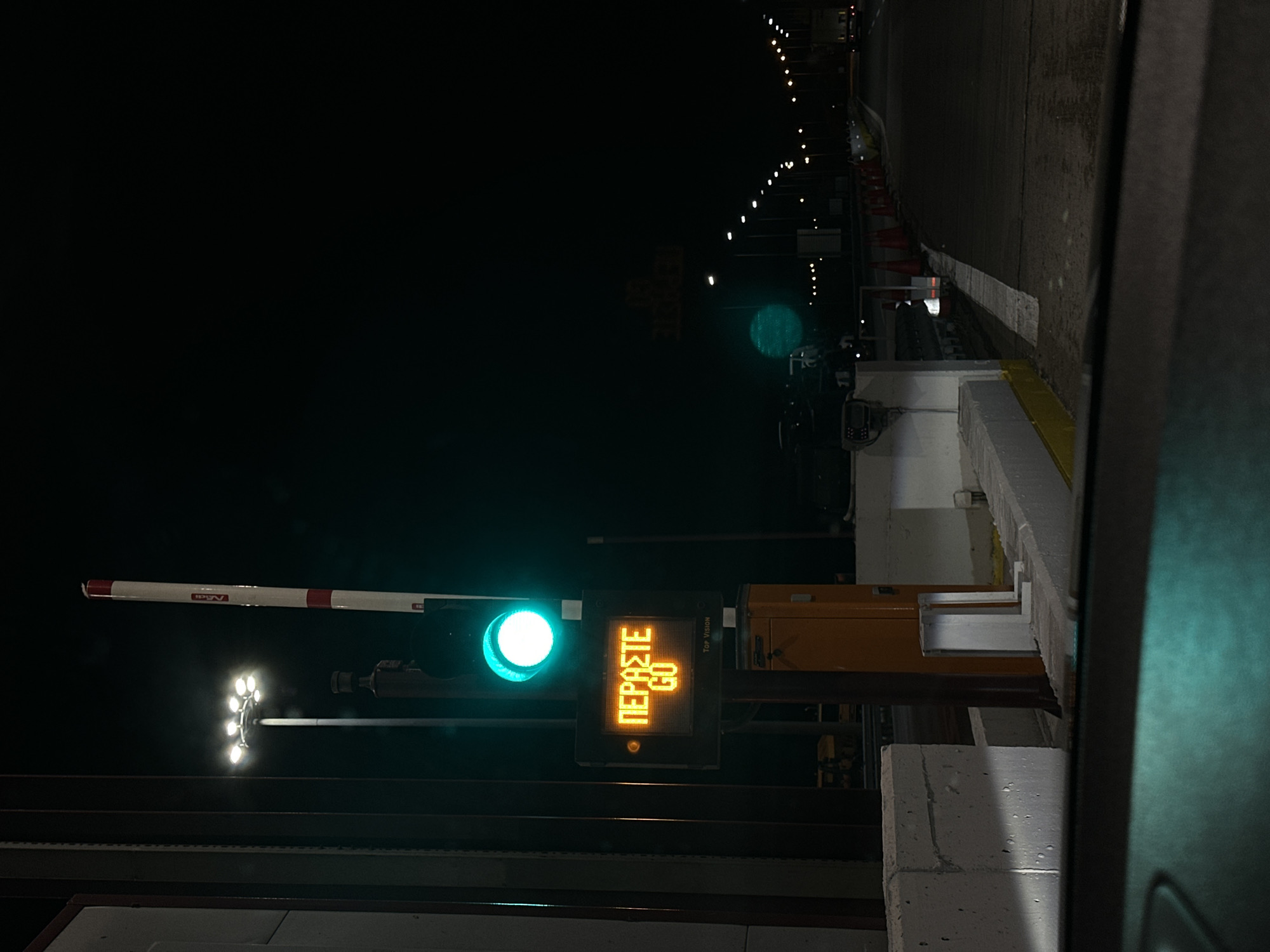 A ground-level, nocturnal shot captures a checkpoint in Δημοτική Ενότητα Προμαχώνος, Greece. A white-and-red gate arm is raised, beside a green traffic light and an electronic sign displaying "ΠΕΡΑΣΤΕ" and "GO." A cluster of spotlights is visible further back. The location appears to be a toll booth with white concrete barriers and a yellow structure. In the distance, a road is lit by streetlights with orange traffic cones. A vehicle's interior is vaguely visible in the right foreground. The scene is largely dark, punctuated by artificial lights.
