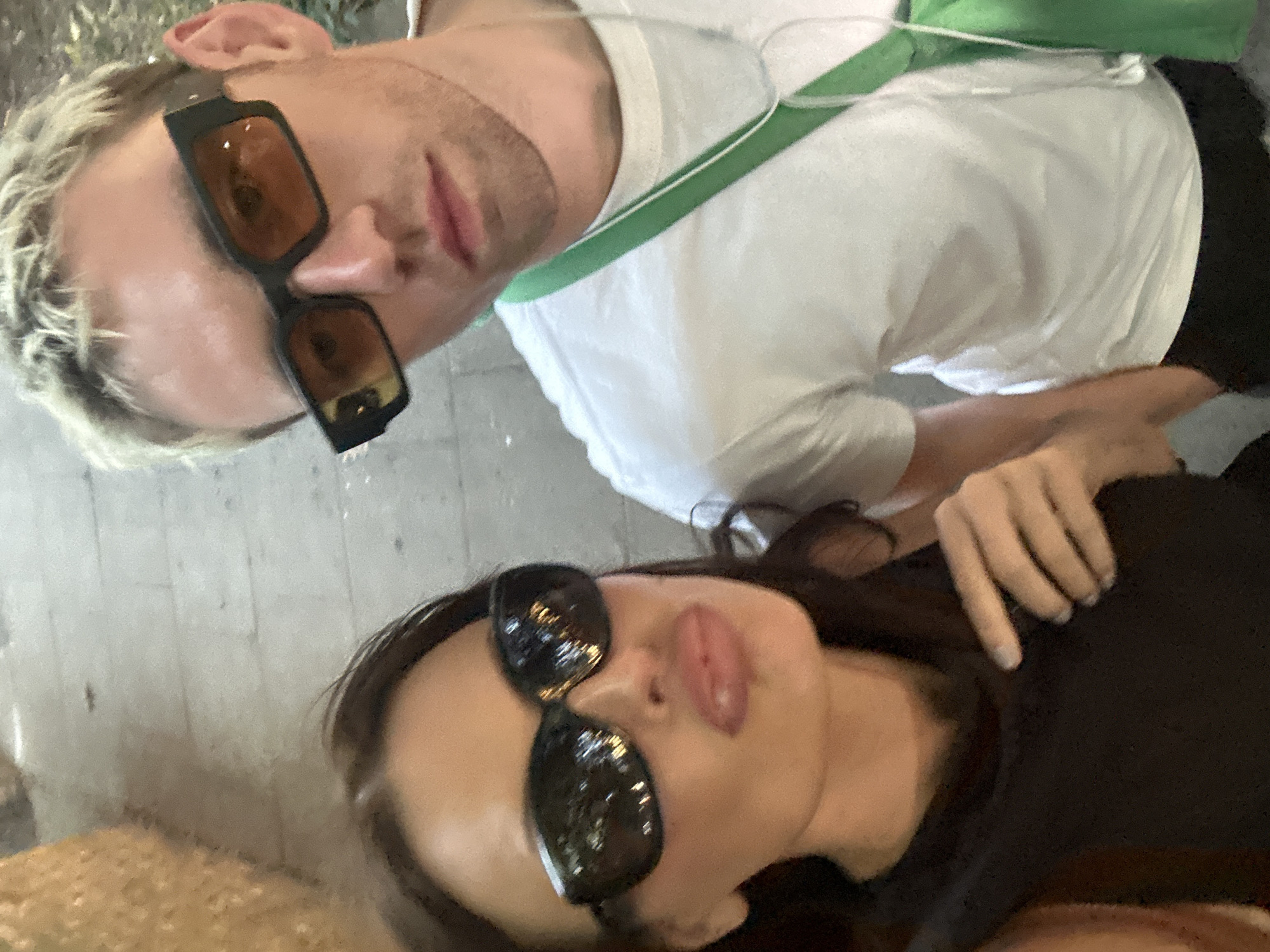 In a candid, close-up, outdoor selfie, two individuals are positioned in the foreground against a blurry urban backdrop that may be a cobblestone street in London, United Kingdom. The person on the left, a woman with dark hair, is wearing large, dark sunglasses and a black top, her lips are pursed. The person on the right, a man with blonde, spiky hair, sports similar large, dark-rimmed sunglasses with amber lenses and a white t-shirt, a bright green strap is visible across his chest. Both individuals are looking towards the camera, suggesting a shared moment captured while walking or exploring. The lighting appears to be natural daylight, with a slightly warm tone.
