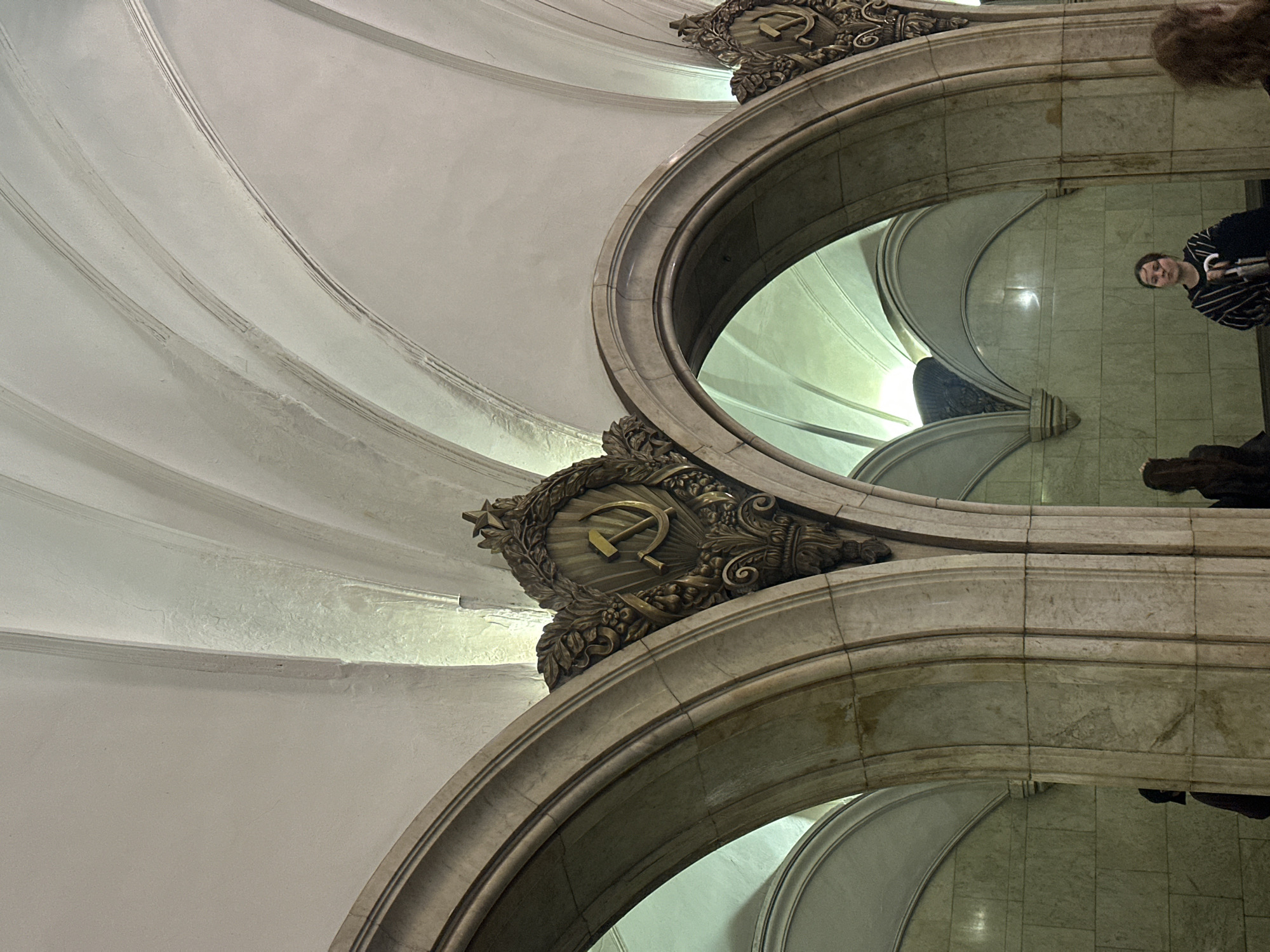 This media file shows an interior architectural detail of a grand, possibly public building in Moscow, Russia. The dominant elements are ornate marble arches and curved, textured walls that create a sense of depth and grandeur.
Prominently featured are Soviet-era symbols. Above one of the arches, a golden emblem of a hammer and sickle within a wreath is visible. This suggests the location might be a historic building, a metro station, or a monument from the Soviet period.
Reflected in the polished marble surfaces are parts of the scene and people. In the upper right reflection, a woman with brown hair, wearing a striped shirt, is visible. She appears to be looking towards the camera. Another reflection shows a darker, indistinct form, possibly another person or a statue.
The lighting is somewhat dim, with artificial light sources illuminating the architectural features, creating shadows and highlighting the textures of the marble and the decorative elements. The overall impression is one of historical significance and architectural splendor. The presence of people indicates that the location is accessible and likely in use. There is no visible text in the image.