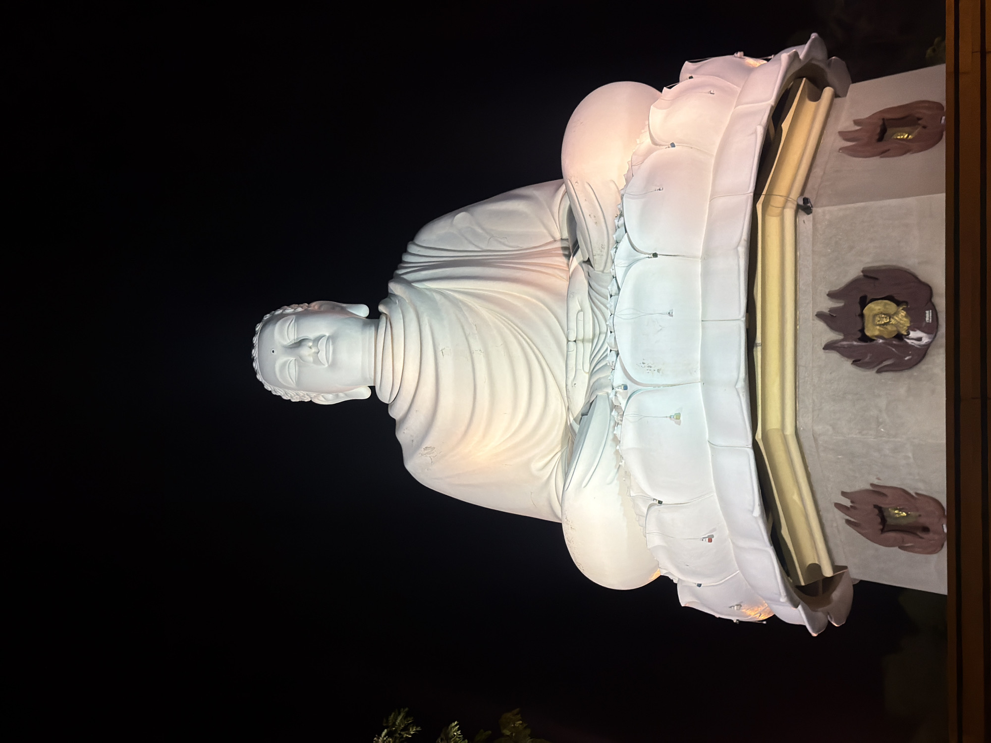 A large, white Buddha statue is illuminated at night, seated on a lotus flower base. The statue is a prominent landmark in Nha Trang, Vietnam. The statue's serene expression and the peaceful posture suggest a religious or spiritual context. The dark background of the night sky contrasts with the bright illumination of the statue, creating a dramatic and awe-inspiring effect. The statue is situated on a tiered pedestal adorned with decorative elements, including flame-like motifs and small golden figures. The overall scene evokes a sense of tranquility and reverence.