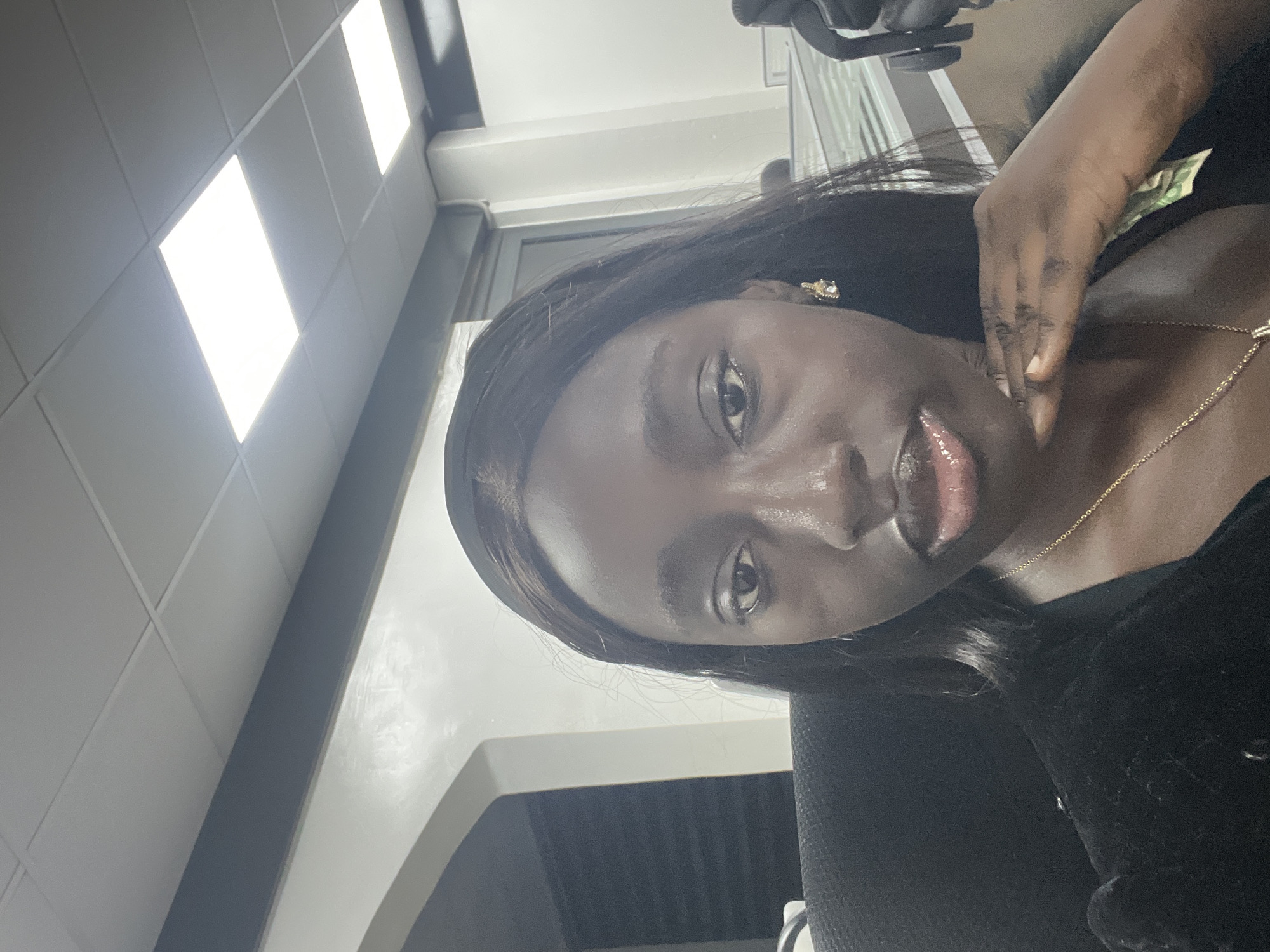 A person is pictured indoors, likely in an office-type setting, based on the ceiling with fluorescent lights. The individual has dark skin, black hair, and is wearing a gold necklace and possibly an earring. Their right hand is touching their neck. The surroundings include a grey wall and what appears to be an office chair in the background. There is no information available to confirm if the location is in Karum, Nigeria.