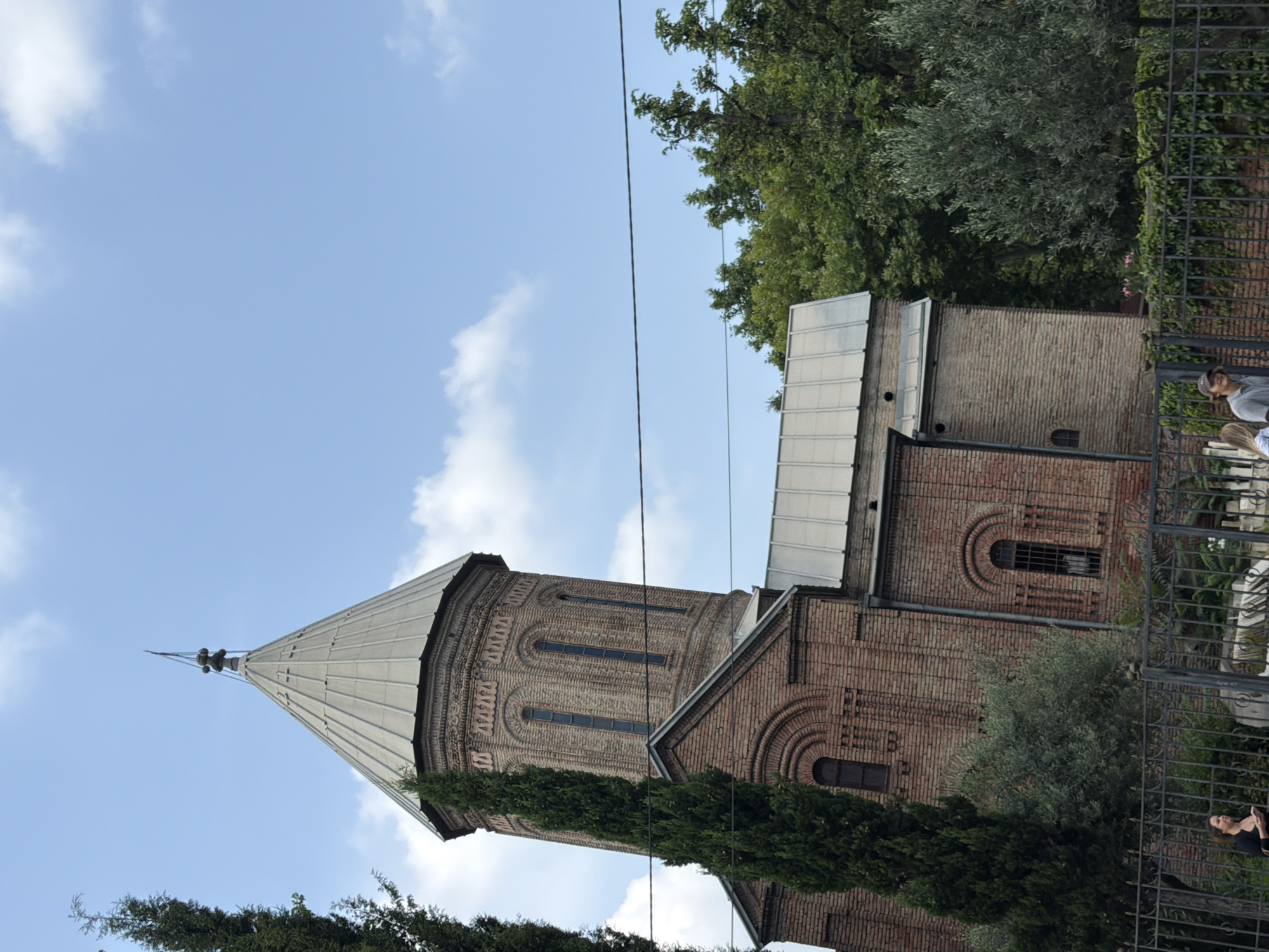 The image shows the exterior of the Holy Trinity Cathedral of Tbilisi, also known as Sameba Cathedral, in Tbilisi, Georgia. The cathedral is constructed of red brick and features a distinctive conical dome topped with a cross. The sky is a clear blue with scattered white clouds. In the foreground, there is a black metal fence, and beyond it, lush green trees and foliage. A few people are visible near the fence, some walking and others standing. The architecture of the cathedral suggests a traditional Eastern Orthodox style. The image appears to be taken during daylight.
