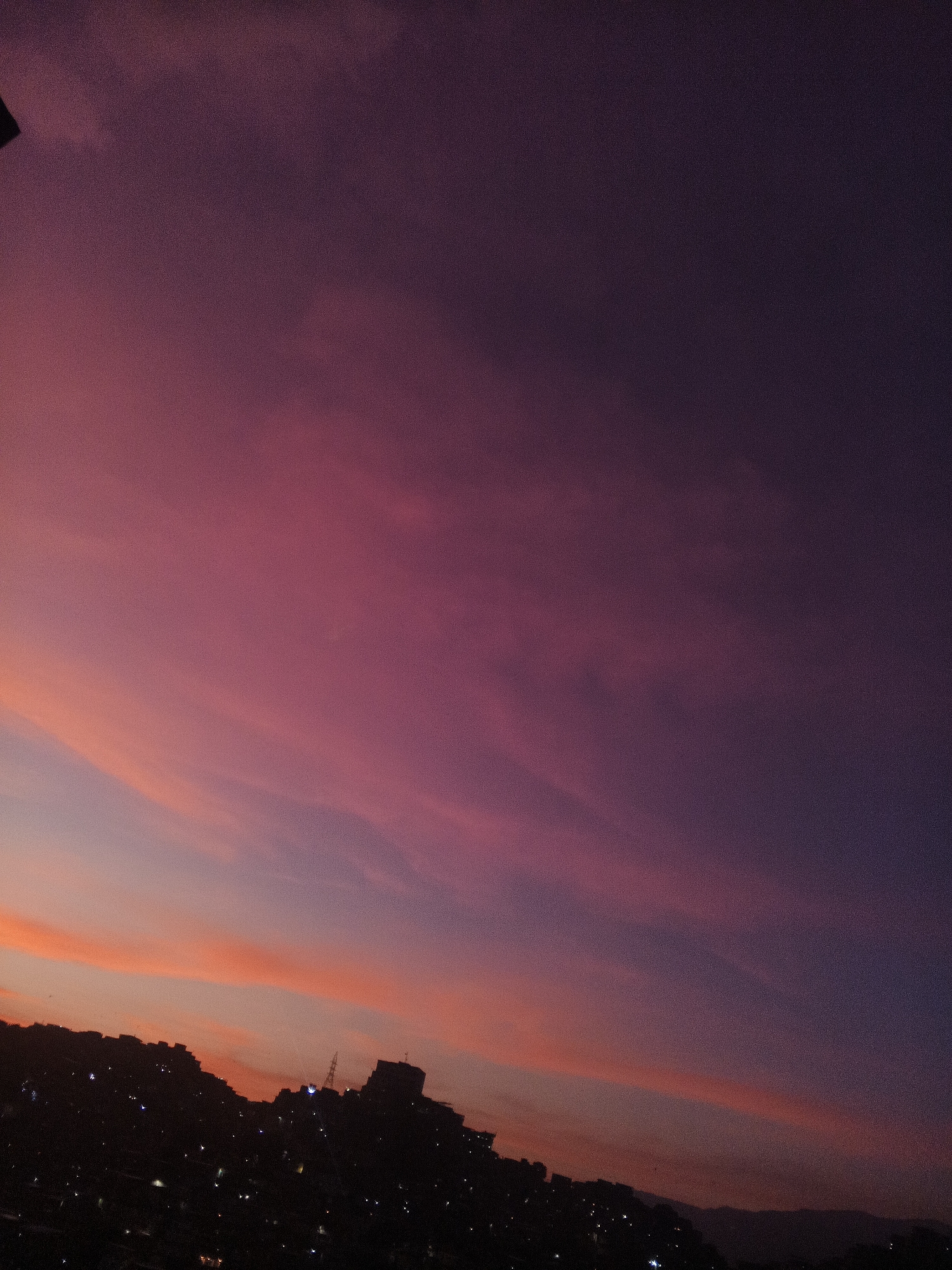A nighttime shot shows a city skyline against a vibrant sunset sky. The sky is primarily shades of deep pink and purple, with lighter orange tones near the horizon. Silhouetted against the colored sky are buildings, some with visible lights, and what appears to be a communications tower. The scene is likely in Caracas, Venezuela. The image primarily captures a twilight cityscape.