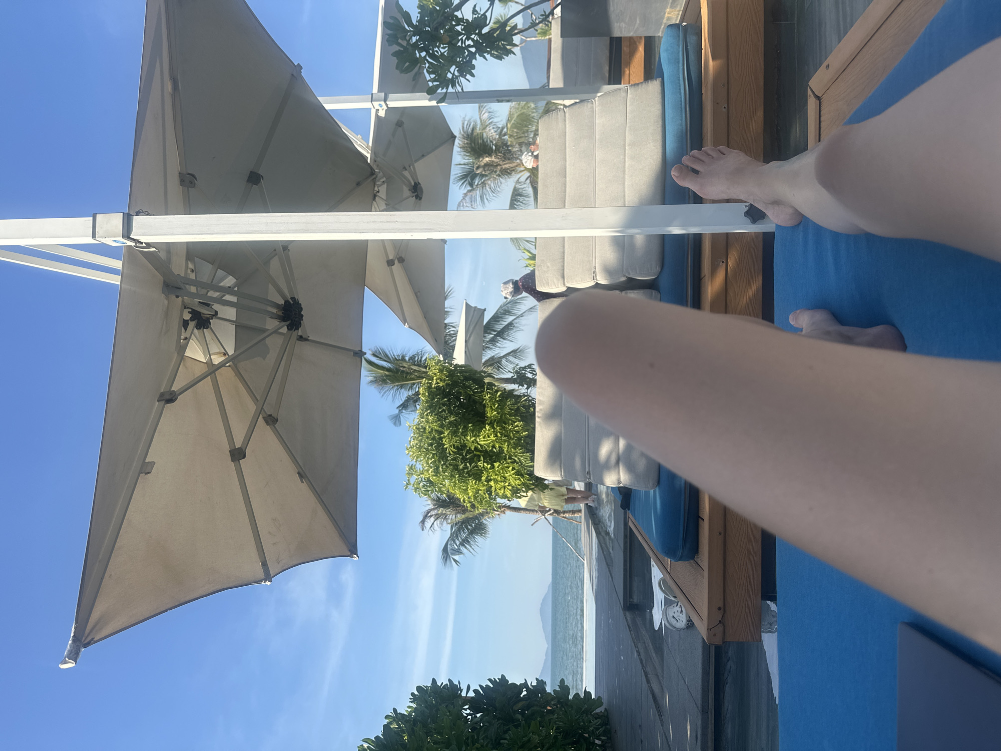 This media file shows a first-person perspective of someone relaxing on a lounge chair by the sea in Da Nang, Vietnam. The scene is set outdoors, under a clear blue sky, suggesting pleasant weather. The view is dominated by large, light-colored patio umbrellas, casting shadows and providing shade. In the background, palm trees and lush green foliage are visible, typical of a tropical or subtropical coastal resort.
The immediate foreground features a person's legs and feet, casually placed on a bright blue cushion, indicating they are likely lounging or sunbathing. A wooden-framed lounge chair with gray cushions is also visible. Further back, the tranquil sea stretches to the horizon, with a hazy mountain range faintly visible in the distance. The overall atmosphere appears relaxed and peaceful, hinting at a vacation or leisure activity. There is no visible text in the media file.