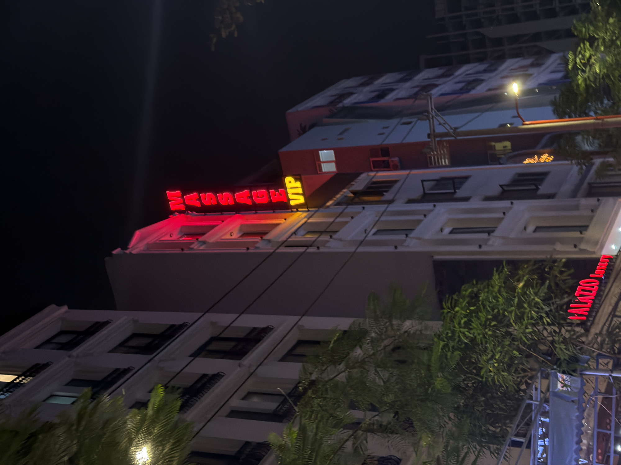 This media file shows a nighttime scene of a building facade in Đà Nẵng, Vietnam. The building appears to be a hotel or a similar establishment, with multiple floors and a modern architectural style. Red neon signs are prominently displayed on the upper floors. One sign reads "MASSAGE VIP" in bright red lettering. Another sign, located further to the right and higher up, reads "PALAZZO Luxury" in red text. The building is illuminated, and there are some lights visible through the windows, suggesting that the interior is occupied. Trees and foliage are visible in the foreground, some of which are illuminated by artificial lights. The overall impression is that of a vibrant urban nightlife scene. There is no clear indication of the weather, but the darkness suggests it is nighttime. The presence of the neon signs and the building's design suggest an entertainment or hospitality venue.