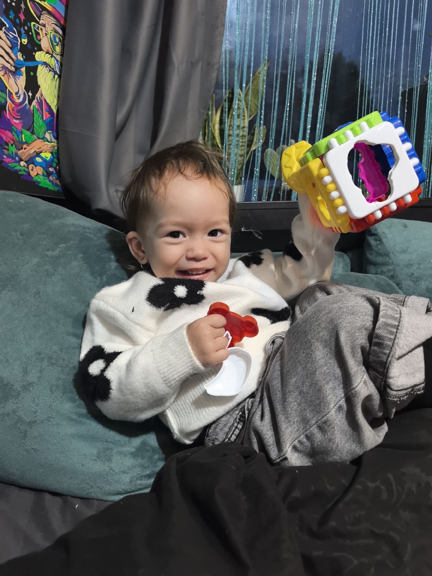 A young child, appearing to be a toddler, is seated on a dark teal upholstered couch, smiling directly at the viewer. The child wears a white sweater with black irregular patterns and grey trousers. A small silver hoop earring is visible in the child's left ear. In the child's right hand, a multi-colored, cube-shaped toy with yellow, green, blue, white, and red components is held; it features a large cutout revealing a magenta interior. The child's left hand holds a small red bear-shaped object and a small white scoop-like object. A dark grey fabric covers a portion of the child's lower body and the couch.
To the left of the child, a brightly colored, neon-style artwork depicting a stylized figure is visible. Behind the child and to the right, a window with vertical blue-teal string or beaded curtains is present. Green foliage is discernible through the window. Dark grey curtains are partially drawn to the left of the window area. The specific location within the city of Noviy, Russia, cannot be determined from the visual evidence provided.