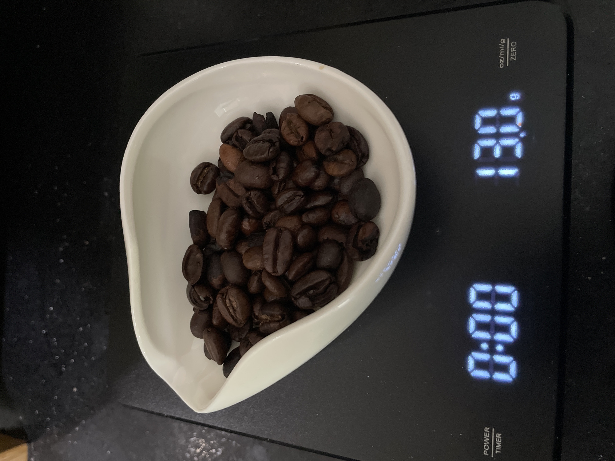The image captures an overhead view of coffee beans being weighed on a digital kitchen scale, set against a dark, speckled background, likely a kitchen countertop. A pile of dark roasted coffee beans rests within a white, irregularly shaped ceramic scoop or shallow bowl. The beans appear rich brown to dark brown, suggesting a medium to dark roast.
The digital scale, black in color, displays two distinct readouts. The upper display shows "13.0 g", indicating a weight of 13.0 grams, with "oz/ml/g" and "ZERO" printed vertically to its left. The lower display shows "00:00", suggesting a timer function that is currently at zero. Below the timer display, the words "POWER" and "TIMER" are printed vertically. The blue glow of the digital numbers contrasts with the black surface of the scale.
The activity depicted is the precise measurement of coffee beans, a common step in preparing coffee. The scene is functional and focuses on the ingredients and tools involved in brewing. No people are visible, and there are no direct cues within the image to indicate the provided location context of Kuta Selatan, Indonesia. The lighting is even, suggesting an indoor setting without specific time of day cues.