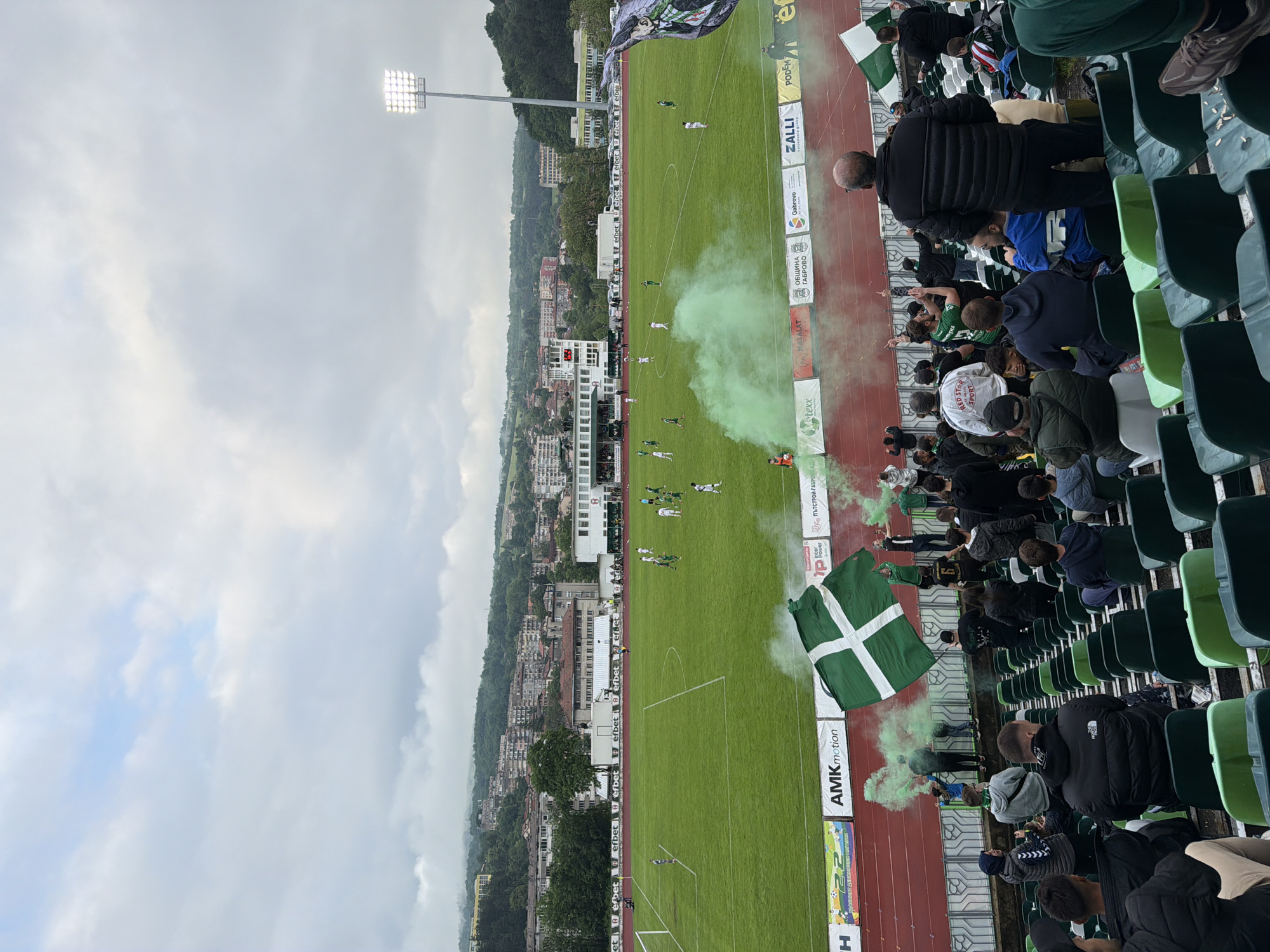 The image captures a soccer game in progress at a stadium, possibly in Gabrovo, Bulgaria based on the visible signage "ОБЩИНА Gabrovo" (Municipality Gabrovo). The game is being played on a bright green field under an overcast sky. A light tower stands in the corner of the field. The city's buildings and forested hills are visible in the background. The stadium is filled with spectators in green and white, holding green and white flags. Green smoke is emitted near the spectator section.
The spectators are seated in the stadium's green seats.
There are advertising boards running along the side of the soccer field. Logos on the boards read "ZALLI", "PODEM", "MAZALAT", "texx", "efbet", "AMKmotion", and "RED STER SPORT."
The stadium's running track is visible in red.