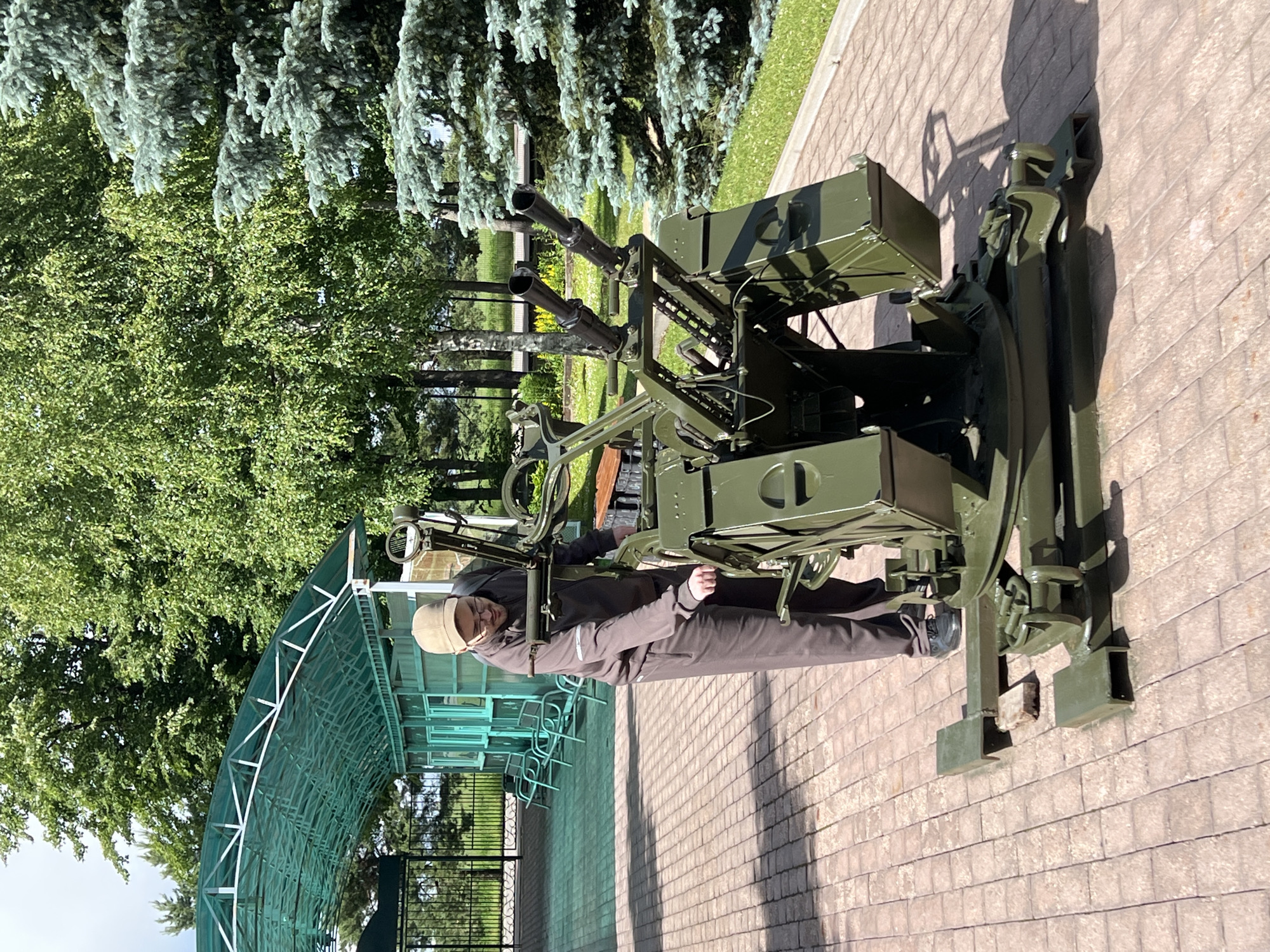 A man wearing a brown tracksuit and a tan baseball cap is standing next to a green anti-aircraft gun on display. The gun has four barrels and is mounted on a wheeled base. The man is reaching towards the gun with his right hand. He is wearing glasses and appears to be looking at the gun. The background shows a paved area with trees and a covered seating area with benches. The scene is outdoors, likely in a museum or park. This is located at the Patriot Congress and Expo Center in Kubinka, Russia.