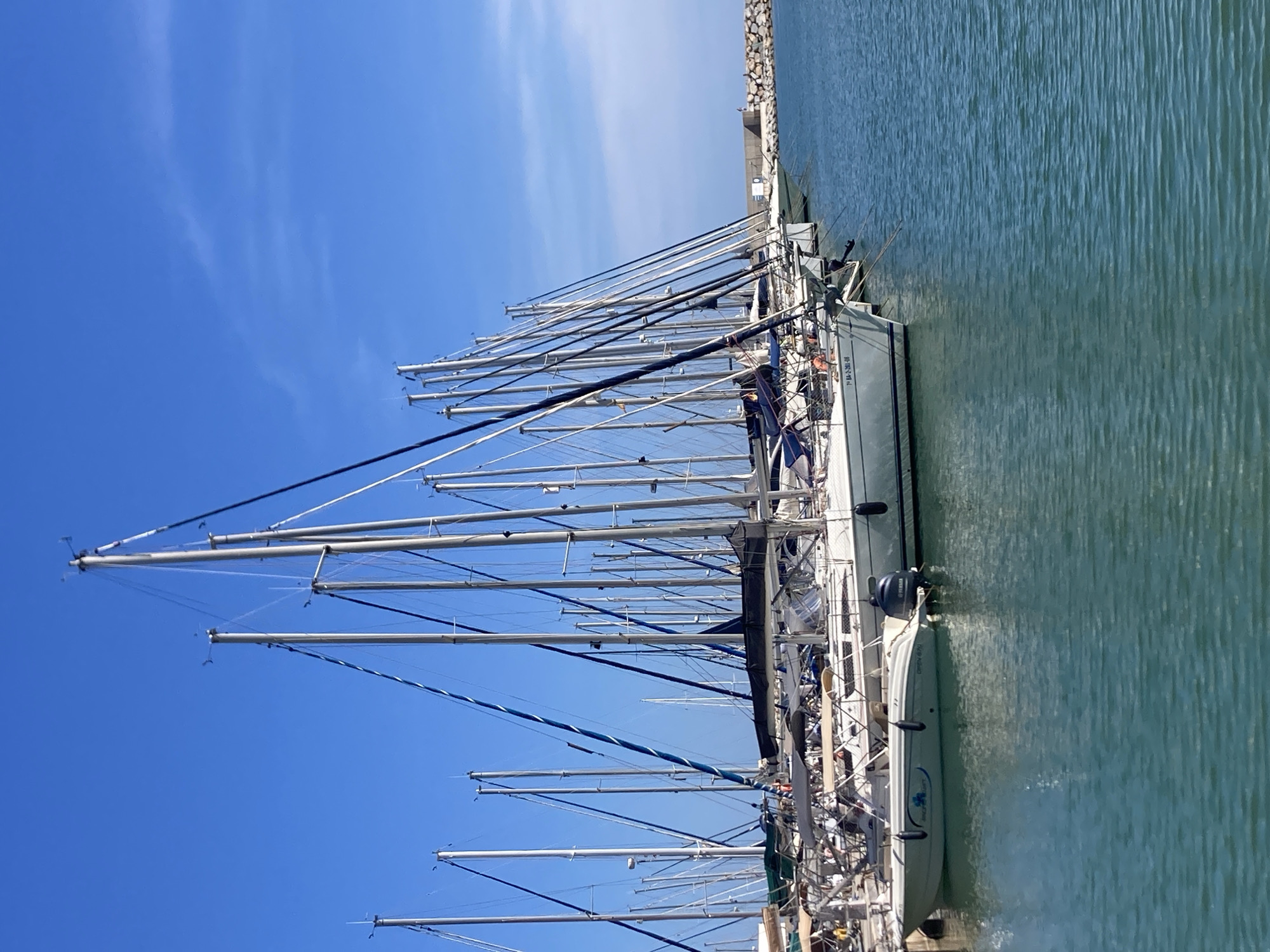 The image shows a marina with numerous sailboats docked on a clear, blue day. The boats are lined up parallel to the viewer, with their masts reaching high into the sky. The water is a calm, turquoise color. In the background, a stone breakwater is visible, and beyond that, a clear blue sky with a few wispy clouds. One of the boats in the foreground is a smaller white motorboat with a Yamaha outboard motor and a logo on its side. Another sailboat in the foreground has the registration number "71 BA-2-261-08" visible on its hull. The location appears to be Port De La Ginesta, Spain, suggested by the style of the boats and the characteristic Mediterranean blue sky. There are no people clearly visible in the image.