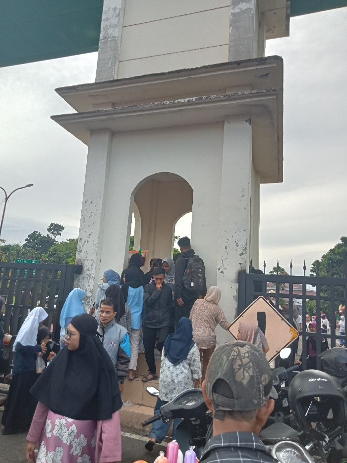 The image depicts a busy outdoor scene in Singaparna, Indonesia, centered around a large, light-colored architectural structure with prominent arches, resembling a gate or a part of a building. The structure shows signs of age with some paint peeling. Overhead, a green, elevated structure, possibly a bridge or overpass, is partially visible.
A crowd of people is gathered around the structure, with many individuals, including women wearing hijabs, moving through or standing near its arched entrance. The attire of the people suggests a casual daytime gathering or transit. In the foreground, a woman wearing a black hijab and a floral-patterned top is visible, along with a man from behind, wearing a plaid shirt and a camouflage cap. Several motorcycles are parked on the right side, with one black helmet clearly showing the brand "Carolus."
The scene is set outdoors under an overcast sky, indicating a cloudy day. A dark metal fence separates the immediate foreground from the background, featuring white text "ST1 M" on its left section. On the right side of the fence, a yellow diamond-shaped warning sign with a black exclamation mark is visible. People appear to be engaged in general activity, suggesting a public area or a point of entry/exit.