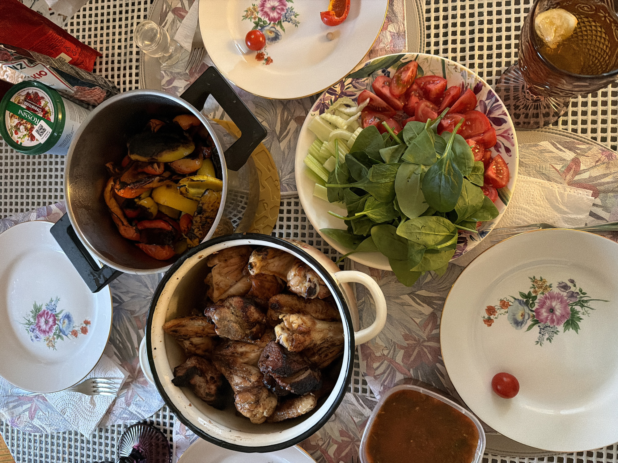 The image captures a dining table laden with an assortment of food items. There are several plates with floral designs, a glass of dark liquid with a lemon wedge, and various pots containing cooked meats, vegetables, and what appears to be grilled peppers. A bowl of chopped tomatoes, celery stalks, and spinach leaves is also visible. Additional details include a can of "ROSSINI" brand food product and other packaged food items. The location is unclear from the image itself, and therefore the city remains Unknown, Unknown.