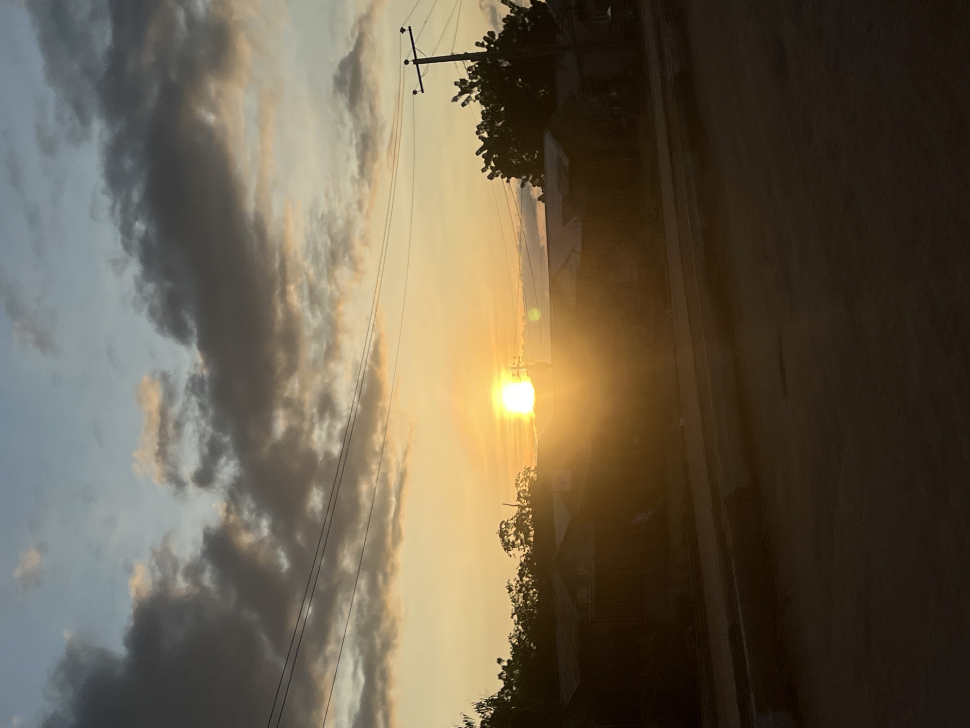 The image displays a sunset or sunrise scene in Howayi, Nigeria, characterized by an intensely bright, circular sun positioned low on the horizon, slightly left of the image's vertical center. Significant horizontal lens flare emanates from the sun. The sky transitions from a light blue-grey at the top, featuring scattered dark grey clouds with some orange illumination, to a vivid orange and yellow closer to the horizon. A prominent, dark silhouette of dense vegetation, likely trees and bushes, spans the midground, obscuring the base of the sun. Below this treeline, several low-rise structures with corrugated metal roofs are visible, also rendered in deep silhouette. Multiple utility lines traverse the upper-mid portion of the sky, and a dark utility pole with horizontal cross arms is present on the far right. The foreground consists of a dark, unpaved ground surface, likely a road, with minimal discernible detail due to deep shadow. No individuals or animals are visible.