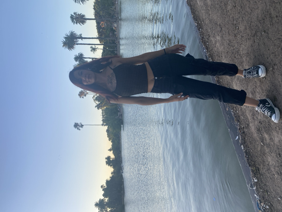 A young woman stands near a body of water, possibly a lake. She has long red hair and is wearing a patterned tank top and dark pants. She also sports dark sneakers. Palm trees are visible in the background, and the sky is a light blue, suggesting it's either dawn or dusk. The scene has a calm, natural ambiance.