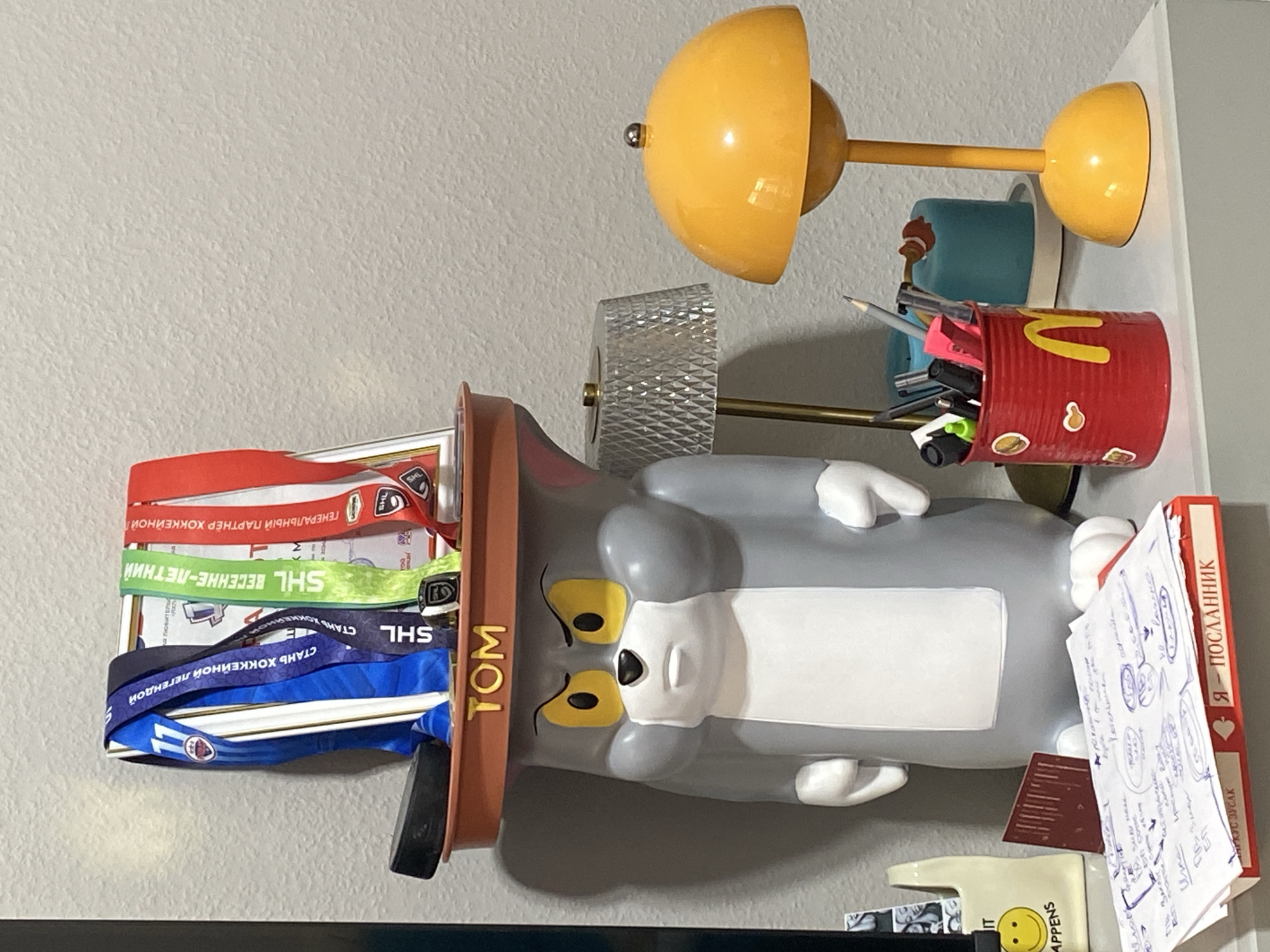 The image depicts a shelf with various objects, including a Tom cat figurine, medals, a hockey puck, a desk lamp, and a pen holder. The figurine is a large, gray, cartoonish representation of Tom the cat wearing a brown hat with the word "TOM" in yellow letters. On the hat, there are several sports medals with ribbons of different colors, including blue, green, and red. A black hockey puck rests on the edge of the hat. To the right of the figurine, a modern-looking desk lamp with a yellow dome-shaped shade is visible, along with a glass lamp with a diamond-patterned shade. A red can, decorated with McDonald's logos and stickers, serves as a pen holder filled with pens and pencils. In the foreground, a stack of papers and a red box with playing card symbols are present. The background appears to be a textured wall. Based on the presence of Cyrillic text on the medals and playing cards, such as "Я — ПОСЛАННИК" (I am an Emissary), and the medals themselves which appear to be related to hockey events with Cyrillic inscriptions like "СТАНЬ ХОККЕЙНОЙ ЛЕГЕНДОЙ" (Become a Hockey Legend) and "ВЕСЕННЕ-ЛЕТНИЙ" (Spring-Summer), it is plausible that this scene is located in Moscow, Russia, as these are common themes and languages in the region.
