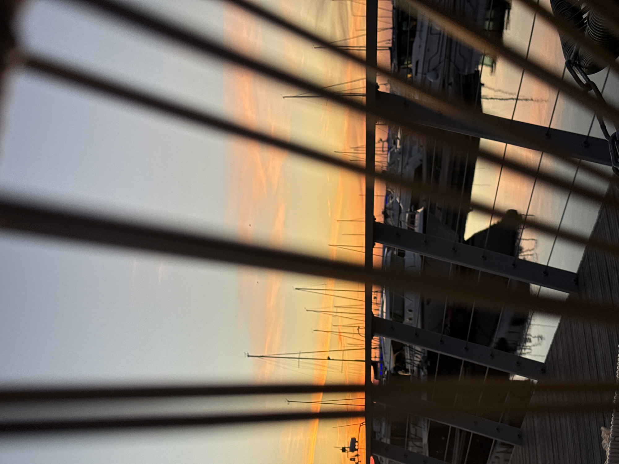 The image depicts a marina at sunset, viewed through the slats of a railing or fence. The sky is painted with warm hues of orange and pink, transitioning into a soft blue at the top. Silhouetted against the colorful sky are the masts of numerous sailboats and yachts. These vessels are moored in the water, which reflects the colors of the sunset. In the foreground, the wooden planks of a walkway are visible, along with parts of the railing system. The overall scene suggests a peaceful evening at a harbor, likely within a coastal city. Given the presence of sailboats and the architectural style of the marina railings, this scene could plausibly be located in Tallinn, Estonia, a city with a notable maritime history and several marinas. However, without specific landmarks or context within the image, pinpointing the exact location to a specific marina in Tallinn is not possible. There are no people visible in the frame. The image is focused on the natural beauty of the sunset and the serene atmosphere of the marina.
