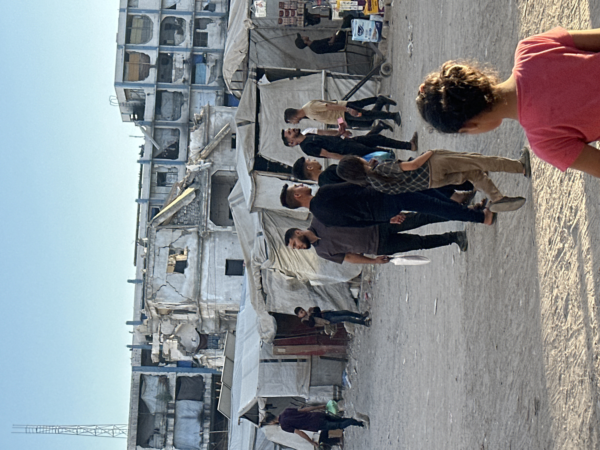 In an urban environment, a group of people are gathered in a dusty, open area. In the background, partially destroyed buildings with exposed rebar and crumbling facades dominate the skyline, suggesting a post-conflict or disaster zone. Several makeshift tents or shelters constructed from tarps are set up in the middle ground.
In the foreground, a young girl with dark, curly hair tied up in a bun, wearing a pink t-shirt, is facing away from the camera. To her left and further back, a group of men and a young boy are walking and standing. One man in a dark t-shirt is walking from left to right carrying a light-colored plastic bag. Another man in a black t-shirt is walking towards the right, followed by a man in a beige shirt. A young boy wearing a vest and jeans stands near one of the tents, looking down. A man with a beard, wearing a dark t-shirt, stands to the right, looking towards the left. A person wearing a black cap is seated near a small stand selling items. The ground is covered in dirt and debris. A tall metal tower is visible on the horizon above the destroyed buildings. The sky is clear and blue.