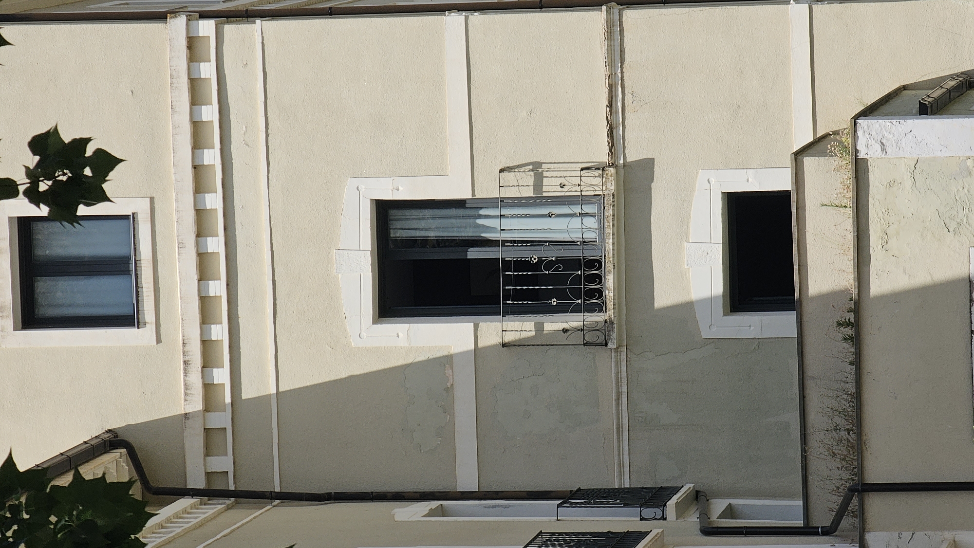 The image shows a facade of a building in Alhama de Aragón, Spain. The building has a light yellow stucco exterior with decorative white trim and molding. There are at least two windows visible. The upper window is a dark frame with light blue curtains visible behind it. Below this is a balcony with an ornate black iron railing. The balcony door is open, revealing a dark interior and light blue curtains. Further down, another window is visible, which appears to be a dark opening. The building is partially shaded by sunlight casting shadows. Some green foliage is visible in the upper left corner, obscuring part of the roofline and gutter. Below the balcony, there is a ledge with some dry vegetation and small green plants growing. The overall scene depicts an architectural detail of a building.