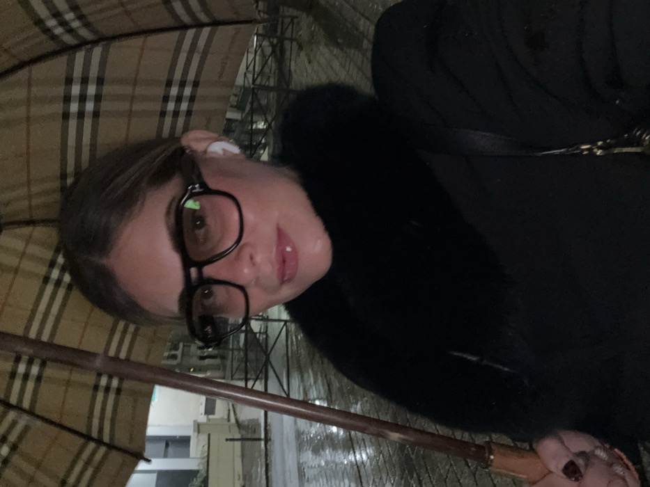 A woman holds a Burberry umbrella in the rain. She wears black-framed glasses and has an earbud in her ear. Her hair is pulled back. She wears a dark coat with fur trim. The street is wet, and buildings are dimly visible in the background.
