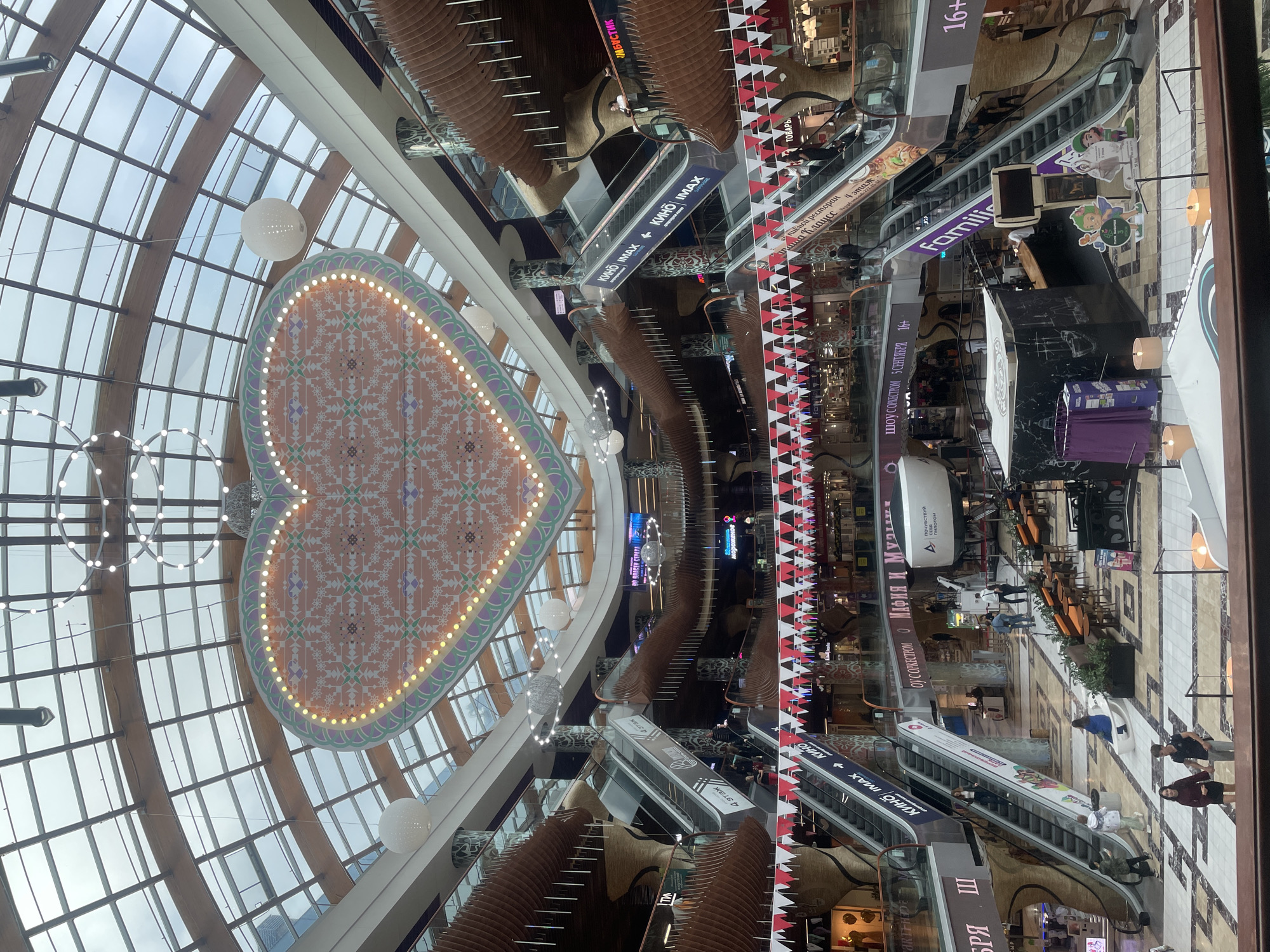 This image shows the interior of the Galleria shopping mall in Saint Petersburg, Russia. The mall features a large, heart-shaped illuminated decoration suspended from the ceiling, which is covered by a glass roof. Multiple levels of escalators and walkways are visible, adorned with red and white triangular flags. People are seen walking on escalators and the ground floor, with some seated at tables in a food court area. The architecture of the mall is characterized by curved wooden elements and patterned pillars. Various signs in Russian are displayed, indicating shops and services within the mall, including "Кино IMAX" (Cinema IMAX) and food establishments.