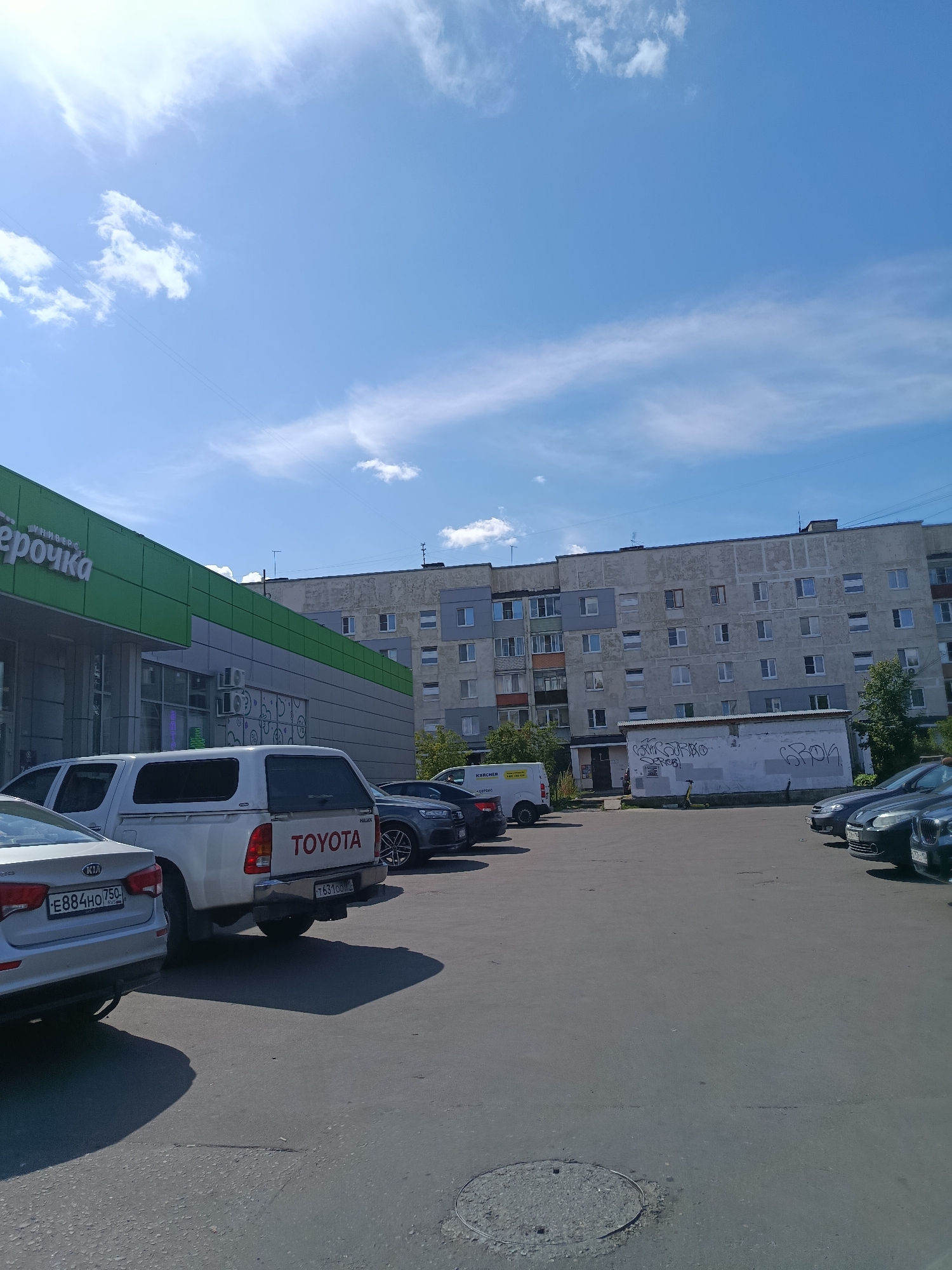 In Orekhovo-Zuyevo, Russia, a sunlit outdoor scene depicts a parking lot in front of a multi-story apartment building and a green-accented commercial building. The sky is blue with scattered white clouds. Several cars are parked in the lot, including a silver Kia, a white Toyota Hilux pickup truck with a camper shell, and various dark-colored sedans. The commercial building, identified by the green signage reading "Pyaterochka," appears to be a grocery store or supermarket. Graffiti is visible on a smaller structure situated between the apartment building and the parking lot. The apartment building is an older, multi-story concrete structure with numerous windows. The overall atmosphere is that of a typical day in a residential and commercial area.