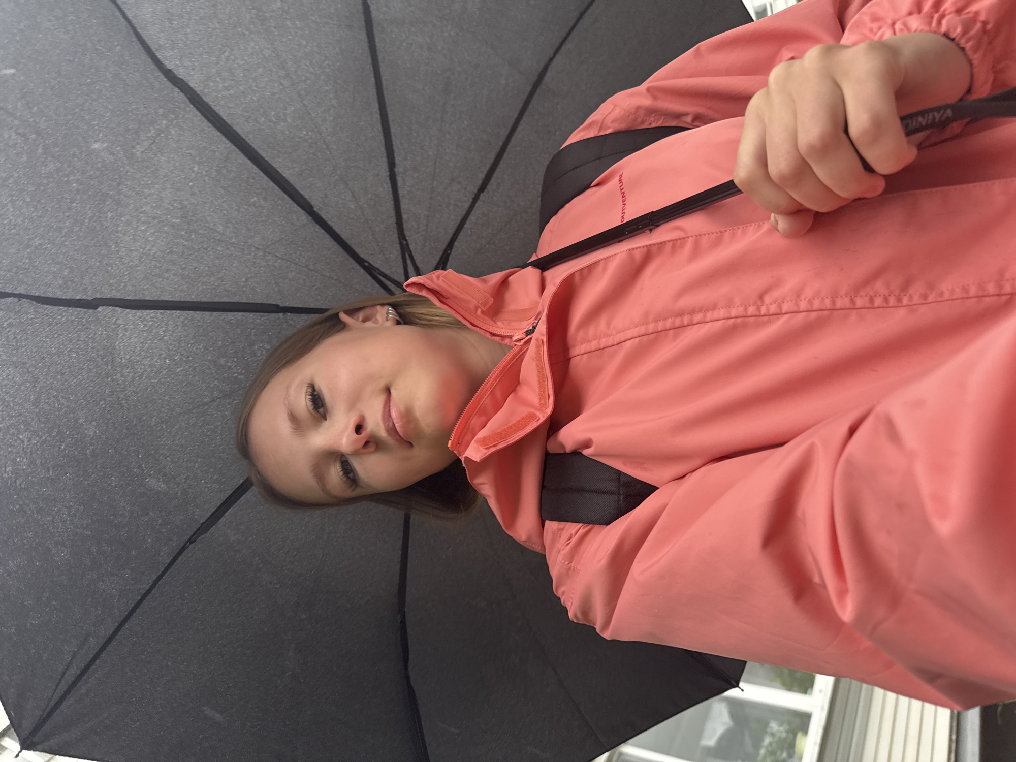 A young girl, likely a teenager, holds a black umbrella over herself as she looks into the camera. She is wearing a bright pink rain jacket with a black backpack underneath. The umbrella is open and angled above her head, casting a shadow. She has blonde hair and is wearing small hoop earrings. The background appears to be an outdoor setting, possibly a residential area, with part of a building and windows visible. The lighting suggests it may be overcast or rainy. The image was taken in Chaikovsky, Russia.