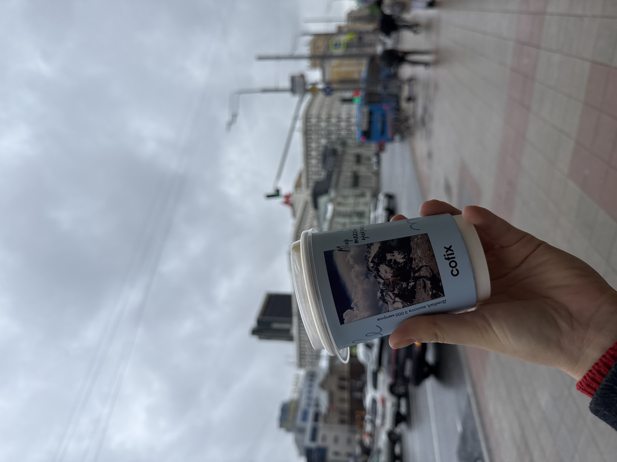 The image, presented with a 90-degree clockwise rotation, depicts a first-person perspective of a hand holding a coffee cup on a city street. The primary subject is a light blue and white disposable coffee cup, held by a person whose hand and a glimpse of a red and dark blue sleeve cuff are visible.
The coffee cup features a printed image of a rugged mountain landscape. Above this image, Cyrillic text reads "Мир может прекрасен" (The world can be beautiful), and below it, "Домбай, высота 3 000 метров" (Dombay, altitude 3,000 meters), referencing a specific mountain region. The brand name "cofix" is also clearly visible in Latin script.
The background, largely out of focus, reveals an urban street scene in Moscow, Russia. Tall, multi-story buildings line the thoroughfare, and elements like traffic lights and power lines are visible. A blue bus and other vehicles are present on the street, and blurred figures of pedestrians can be seen on the sidewalk.
The sky is overcast and grey, indicating a cloudy or dull day. The scene captures a casual, everyday moment of someone holding a coffee cup while presumably walking or standing on a busy city street.