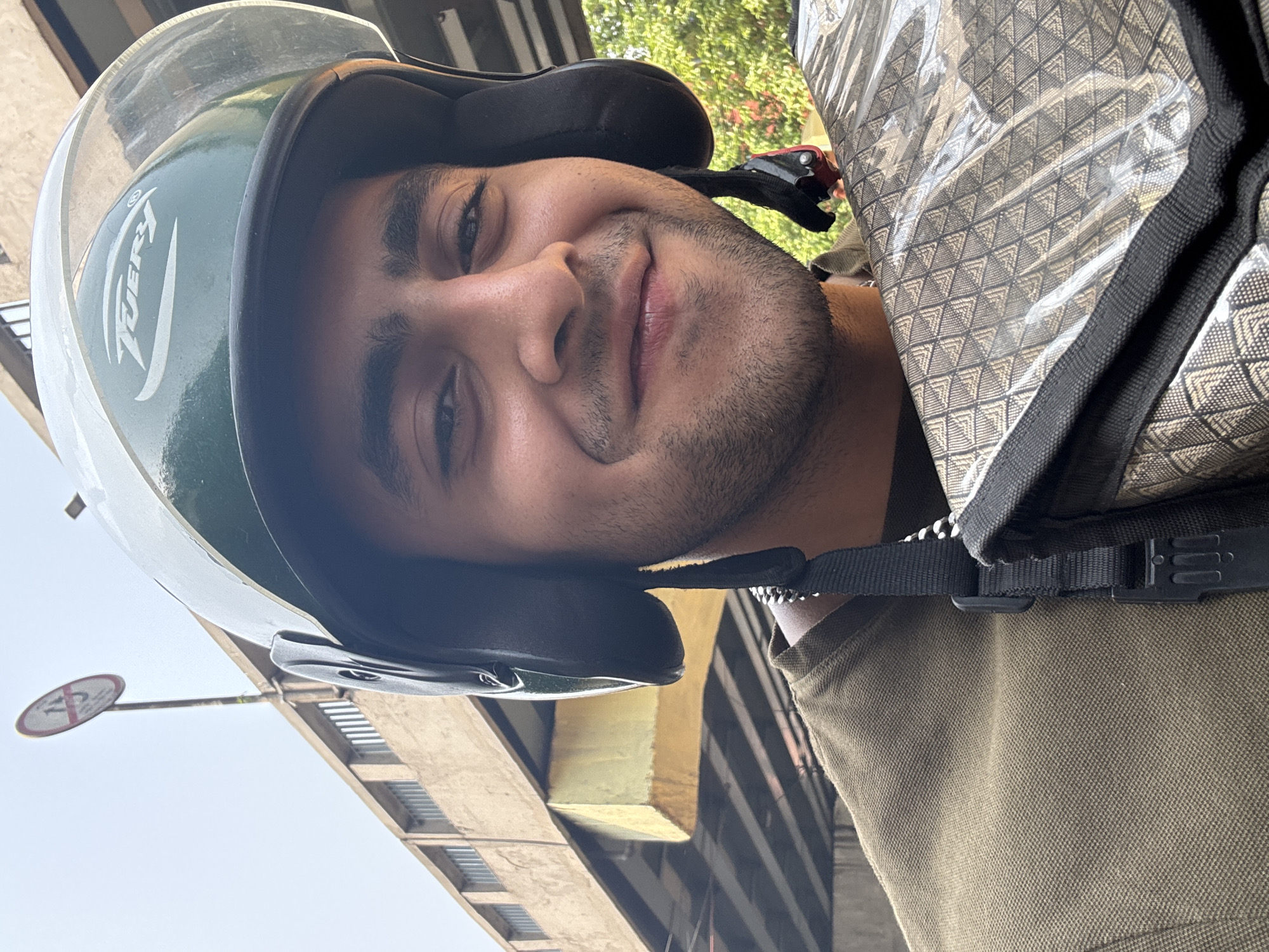 A young man is wearing a helmet and backpack, presumably riding a motorcycle. The helmet is white and green with the word "IVERY" on it. The backpack is gray with a geometric pattern. He has dark hair and a short beard, and is smiling slightly. In the background, there are buildings, one of which is yellow. A "No Entry" sign is visible on a pole. The image was taken in Kolkata, India.