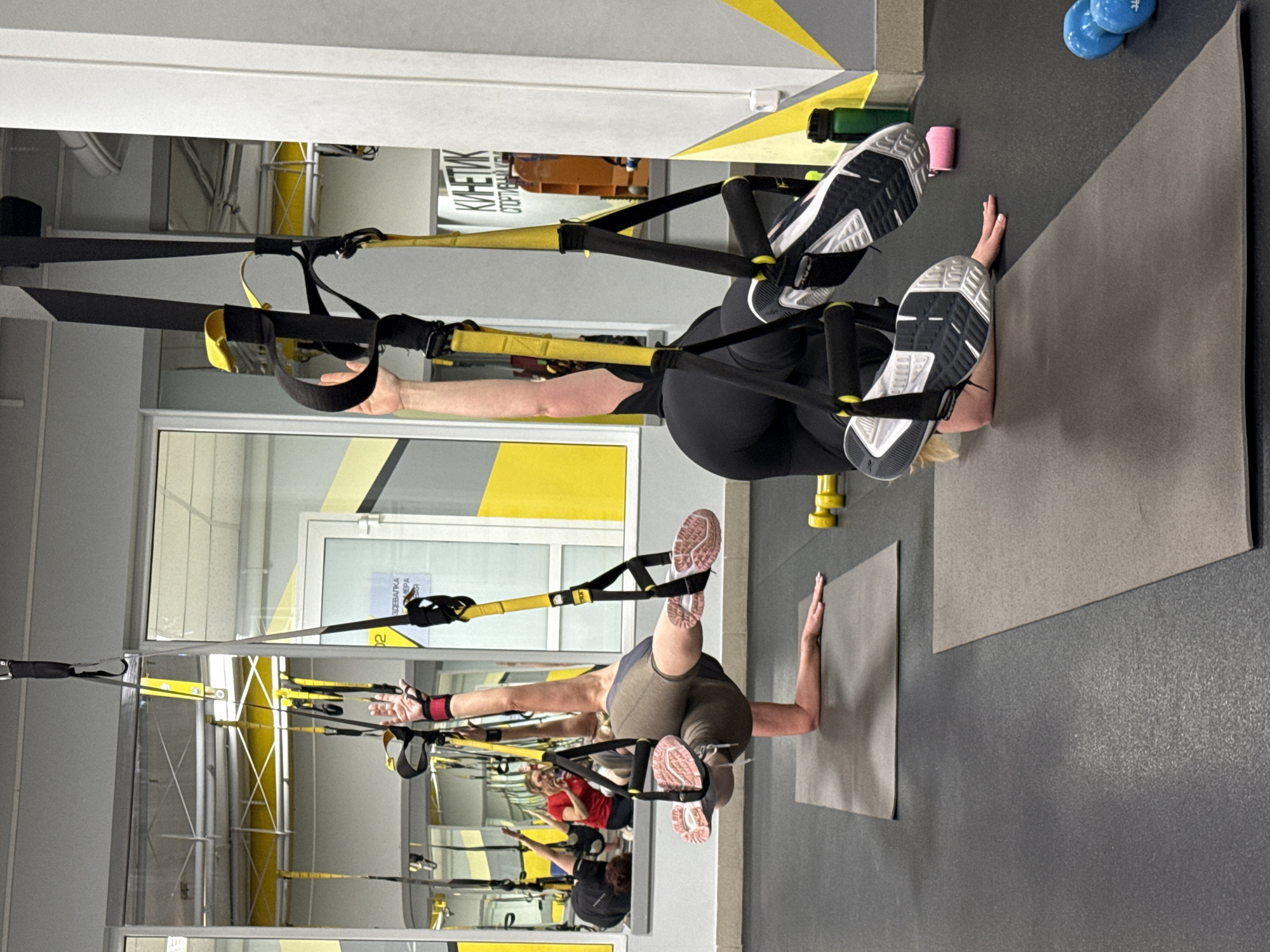 Two individuals are engaged in TRX suspension training within a gym in Chaikovsky, Russia. The setting includes gray and yellow accents, with exercise mats on the floor. One person, wearing black activewear and sneakers, is performing an exercise with their feet suspended in the TRX straps. The second individual, in olive green shorts and athletic shoes, is similarly using the TRX system for another exercise. The gym environment is visible through the window, and the reflections in the mirror provide additional details of the surroundings.