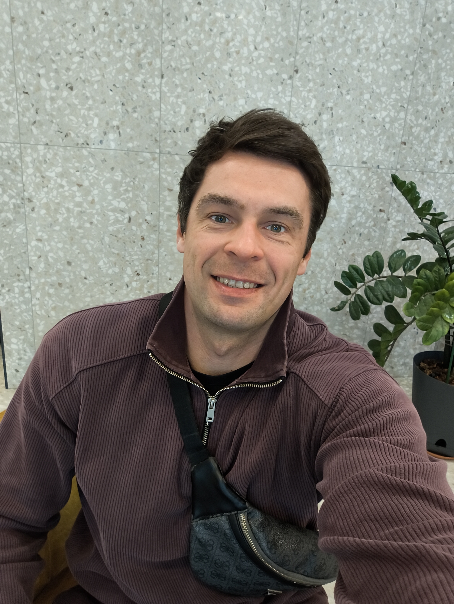 A medium shot presents a man seated indoors, his expression amiable with a broad smile. He sports dark brown hair, and his attire consists of a textured, dark purple, zip-up jacket over a dark under-shirt. A small patterned black bag hangs across his torso.
The backdrop features a neutral-toned wall speckled with small, irregular patterns and a potted plant with dark green leaves on the right.
There is no indication in the image that this was taken in the city of Moscow, Russia.