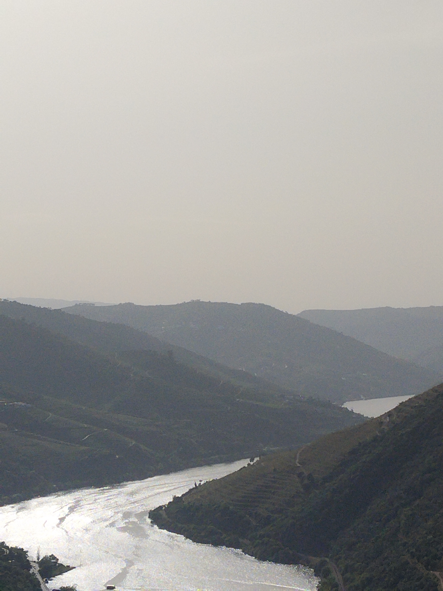 This is a landscape photograph of the Douro River Valley near Tabuaço, Portugal, on an overcast day. The image is taken from an elevated perspective, capturing a wide view of the river winding through a series of rolling hills and terraced vineyards. The river reflects sunlight, appearing as a wide band of shimmering white. The hills are covered in vegetation, and the distinct terraced vineyards are visible on the slopes. In the distance, additional hills fade into the hazy horizon.