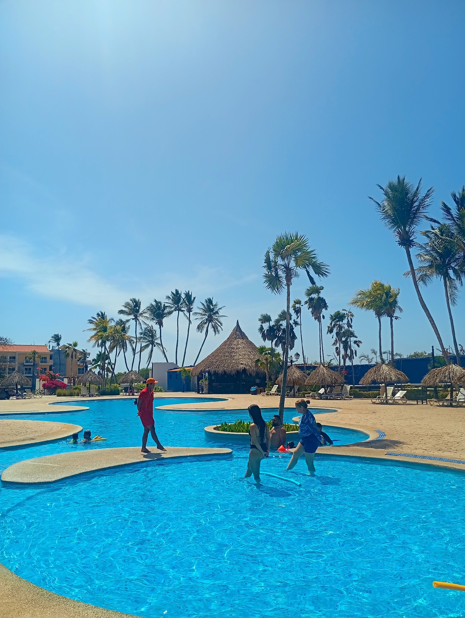 The image depicts a bright, sunny day at a resort swimming pool in Playa El Agua, Venezuela. The pool is turquoise blue and irregularly shaped, with sand-colored islands connected by walkways. Several people are present: a lifeguard dressed in red walks across a pathway, and groups of people are wading in the water. The surroundings feature palm trees, thatched-roof structures providing shade, and lounge chairs. A multi-story building is visible in the background, suggesting a hotel or resort complex.