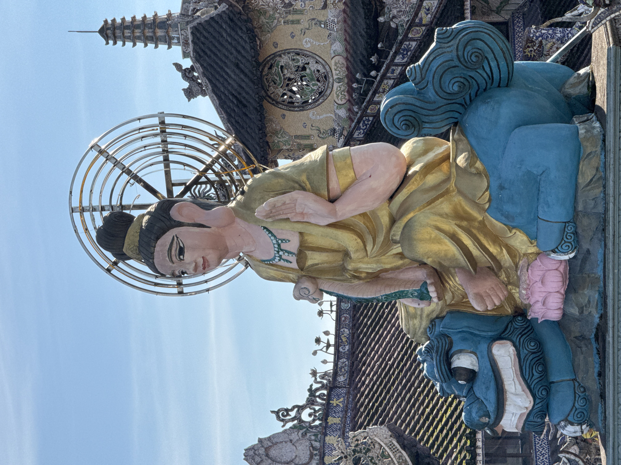 A close-up, medium shot captures a colorful statue of a seated figure, likely a deity or religious icon, with its right hand raised in a gesture of blessing or protection. The statue is adorned in golden robes and a turquoise necklace. It sits atop a bright blue, fantastical creature with a wide, toothy grin and intricate black swirl patterns. The creature rests on a dark, stone-like base. Behind the statue, a traditional Vietnamese temple structure is visible, characterized by dark, tiled roofs and ornate, mosaic-like decorations featuring floral motifs and mythical creatures. A tall, multi-tiered pagoda stands prominently against a clear blue sky. The statue is illuminated by natural sunlight, casting subtle shadows. The scene is likely situated at the Linh Phuoc Pagoda in Da Lat, although the prompt specifies Xuan Thanh, Vietnam.