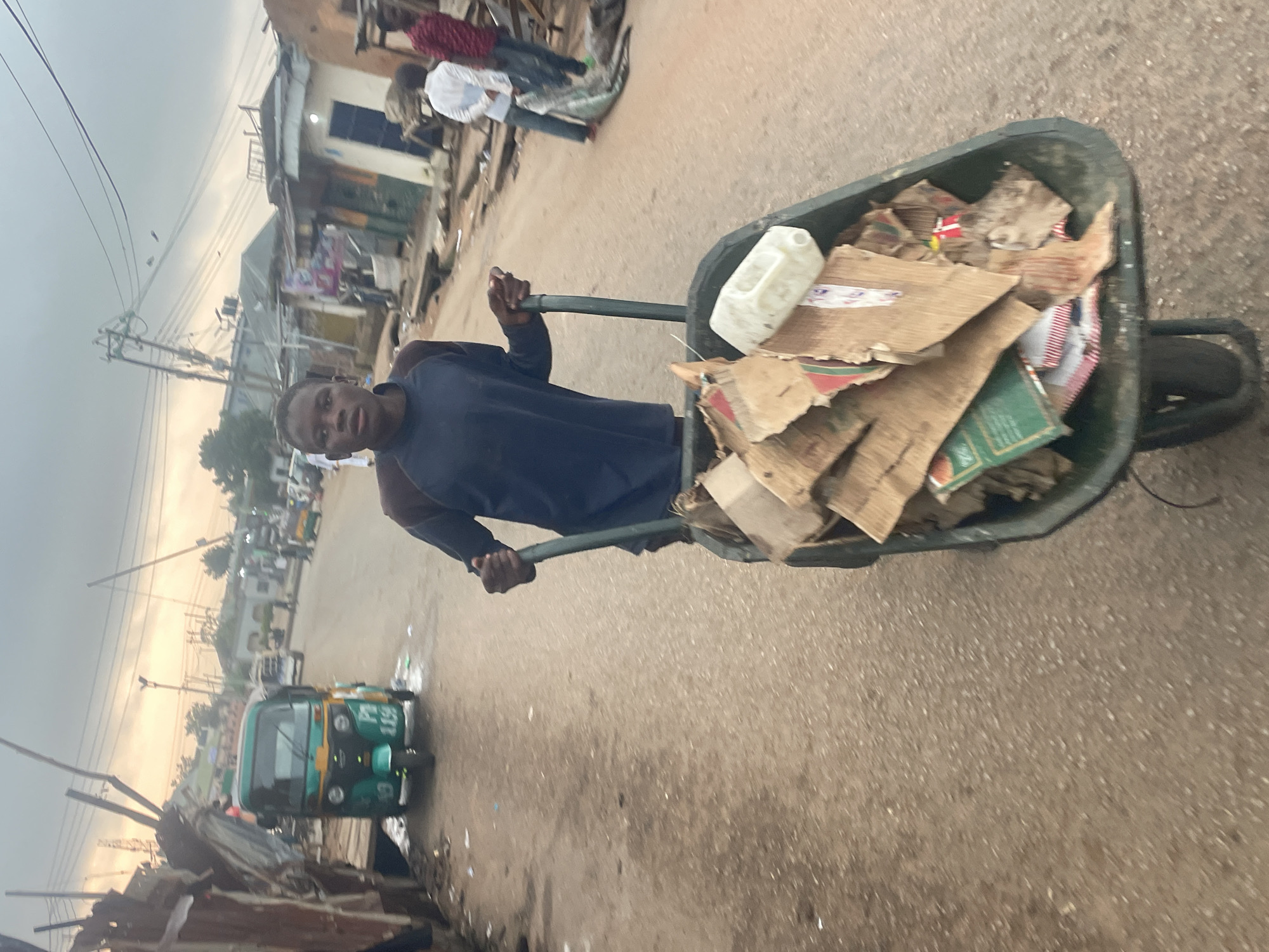 A young person, likely a boy, is pushing a wheelbarrow filled with cardboard and a white plastic container down a dirt road in Gura Top, Nigeria. The person is wearing a dark long-sleeved shirt. The environment appears to be a street scene with some buildings lining the road. In the background, a green and yellow auto-rickshaw is parked, and several other individuals are visible, some standing and others appearing to be walking. The sky is overcast. The road is unpaved and appears somewhat dusty, with debris scattered along the sides.