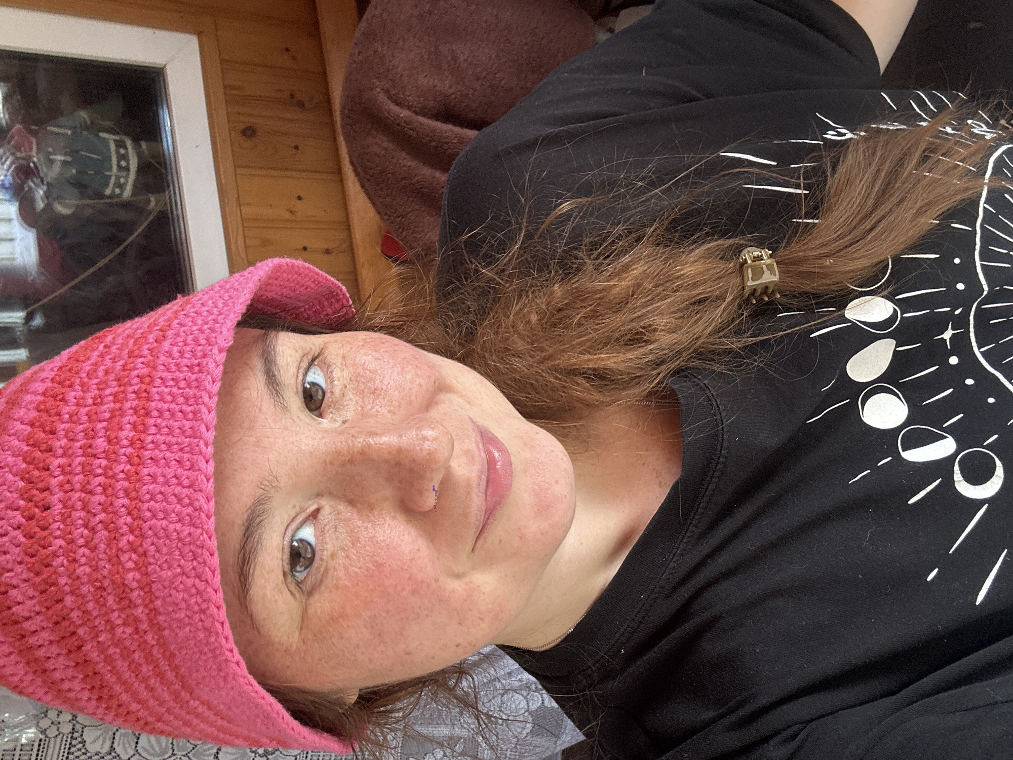A woman with light brown hair, wearing a pink knitted hat and a black t-shirt with a celestial print, poses for a selfie. Her face is visible, showing a nose ring, freckles, and rosy cheeks. She is indoors, with a wooden wall and a window reflecting an interior scene. A brown blanket is visible behind her. The image does not provide any specific clues about the location within the city of Nizhny Novgorod, Russia.