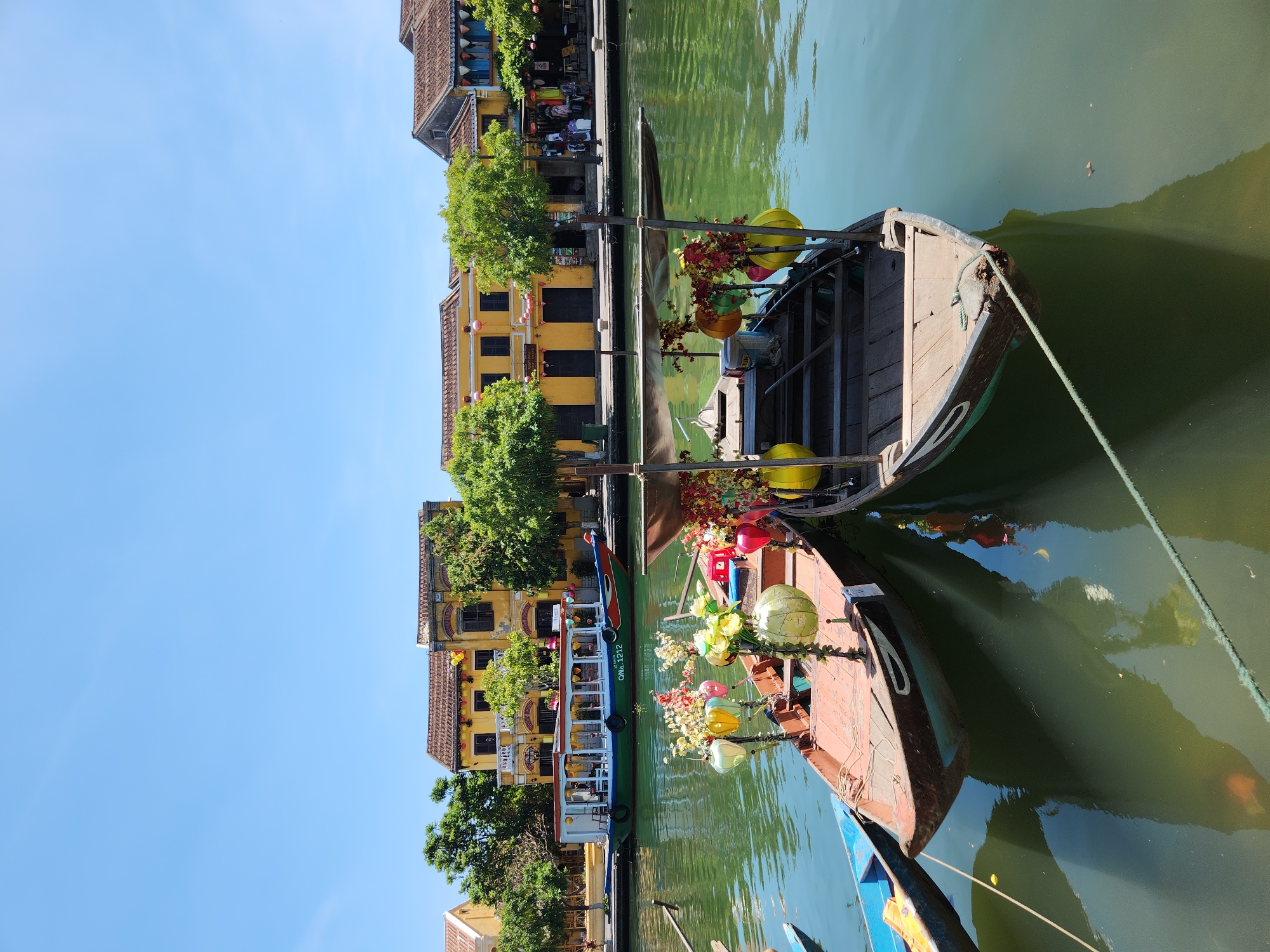 In Hội An Ward, Vietnam, on a bright, clear day, two wooden boats are docked on a calm, green river. The boat in the foreground is reddish-brown and decorated with colorful lanterns and floral arrangements. Behind it, another boat, also wooden, is equipped with a canopy. Both boats are tied with ropes. In the background, traditional yellow-hued buildings with tiled roofs line the riverbank, with lush green trees interspersed among them. A larger, multi-colored passenger boat is visible further down the river. People are seen on the sidewalk in front of the buildings. The scene depicts a serene riverside view of the city.