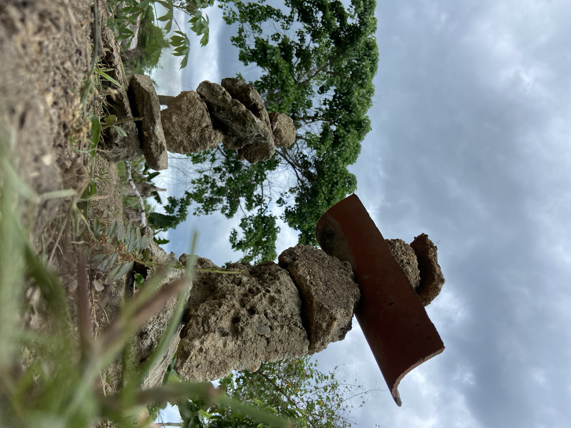 The image depicts two distinct cairns, or stacks of irregularly shaped, rough-textured stones and concrete fragments, situated outdoors in Los Velásquez, Venezuela. In the foreground, dry, light brown soil with sparse green grass and other small plants is visible, with some vegetation significantly blurred due to a shallow depth of field and low camera angle. The cairn on the right is taller and features a reddish-brown, curved object, resembling a broken terracotta roof tile or rusted metal trough, resting atop its uppermost stones, extending diagonally upwards and to the right. The cairn on the left is shorter, composed of similar rough materials. In the mid-ground and background, dense green foliage from large trees, specifically a prominent tree with numerous leaves, is visible. The sky above is overcast, filled with light grey, diffuse clouds. No individuals or animals are present.