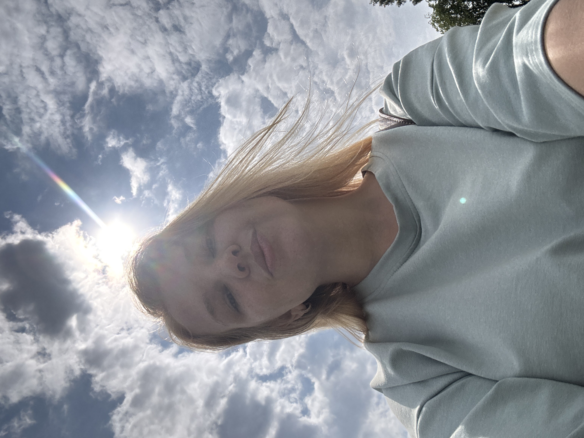 In the city of Breytovo, Russia, a young woman with blonde hair is captured in a selfie outdoors. She is wearing a light blue sweatshirt and looking towards the camera with a slight smile. The background is dominated by a bright, cloudy sky with the sun shining prominently, creating lens flare and a subtle rainbow effect. The clouds are scattered and appear to be in motion, suggesting a windy day. The woman's hair is also being blown by the wind. The image is taken from a low angle, emphasizing the sky and the woman's face. A small portion of a tree can be seen on the right side of the frame.