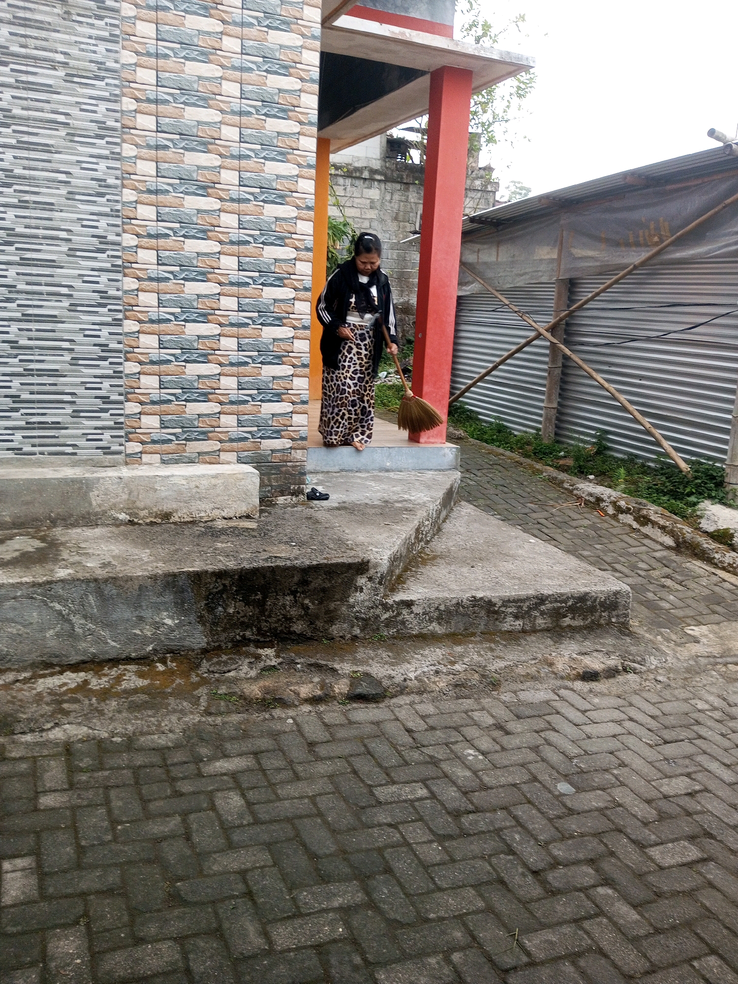 A woman is sweeping a paved area in front of a building. The building has a facade of rectangular tiles in shades of beige, grey, and blue. A red pillar supports a section of the building's roof. To the right of the building, there is a corrugated metal structure covered with a translucent tarp, supported by bamboo poles. The paved area is made of dark grey bricks laid in a herringbone pattern. Concrete steps are present in the foreground. The woman is wearing a black jacket with white stripes and a patterned skirt, and she is holding a broom with a natural fiber head. Her hair is tied back. The scene appears to be outdoors, possibly in a residential or commercial area. The location is Andongsili, Indonesia.