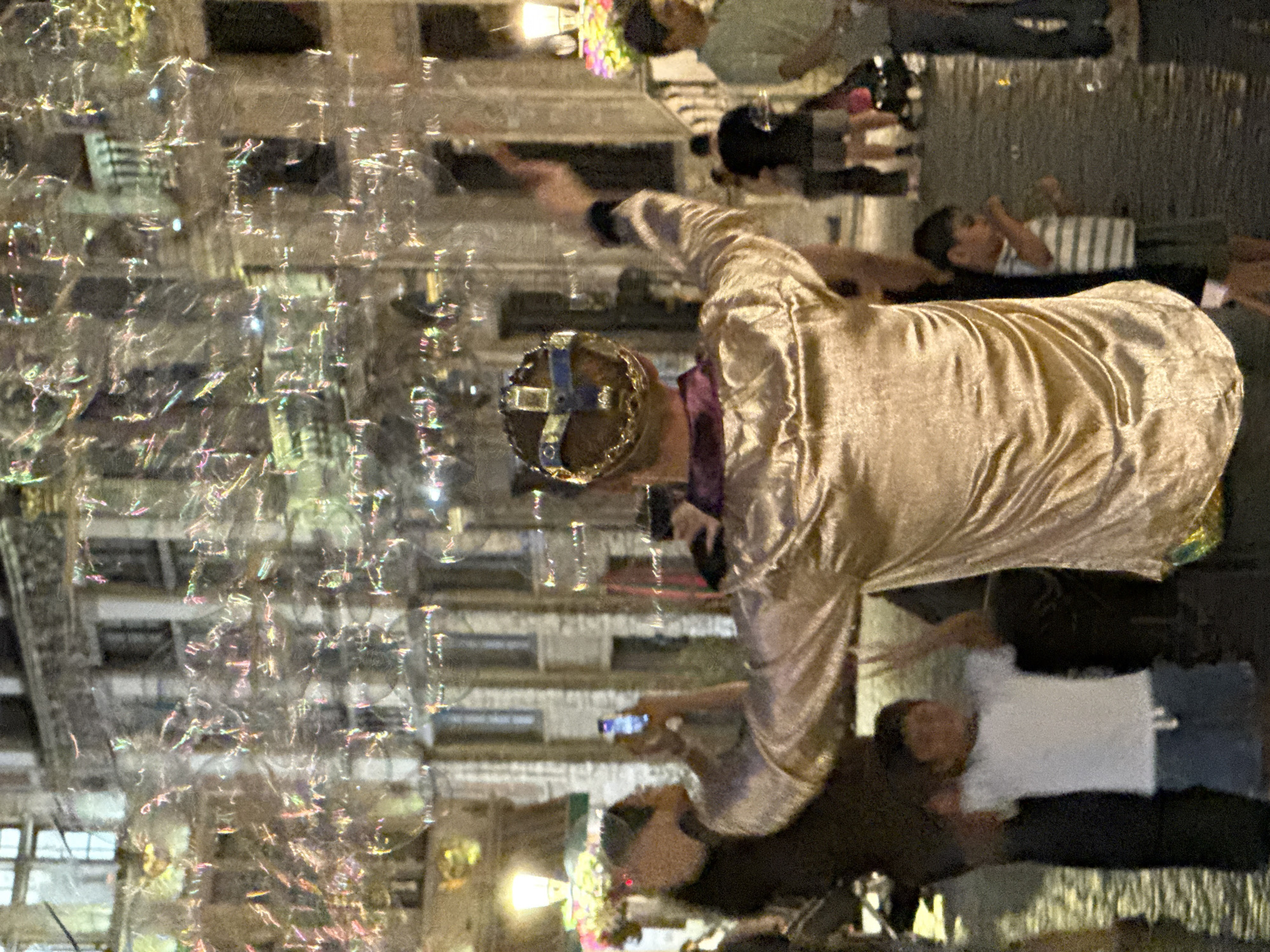 The image depicts a street scene at night in Brussels, Belgium, likely in a tourist area given the architecture and the presence of a street performer. In the foreground, a person wearing a golden, shimmering, long-sleeved shirt and a jeweled crown is blowing bubbles. Their arms are raised, and bubbles are visible floating in the air, reflecting the ambient light. To the left of the performer, a young child in a white t-shirt and blue shorts with their hands raised stands near a darker figure. To the right, a child in a striped shirt and dark shorts is visible. Several other individuals are blurred in the background, along with illuminated buildings featuring ornate architectural details and windows, suggesting a historic European city. The street surface appears wet, possibly from recent rain, reflecting the lights and creating a somewhat blurred and atmospheric effect. The overall scene captures a moment of festive activity, likely an outdoor performance or celebration.