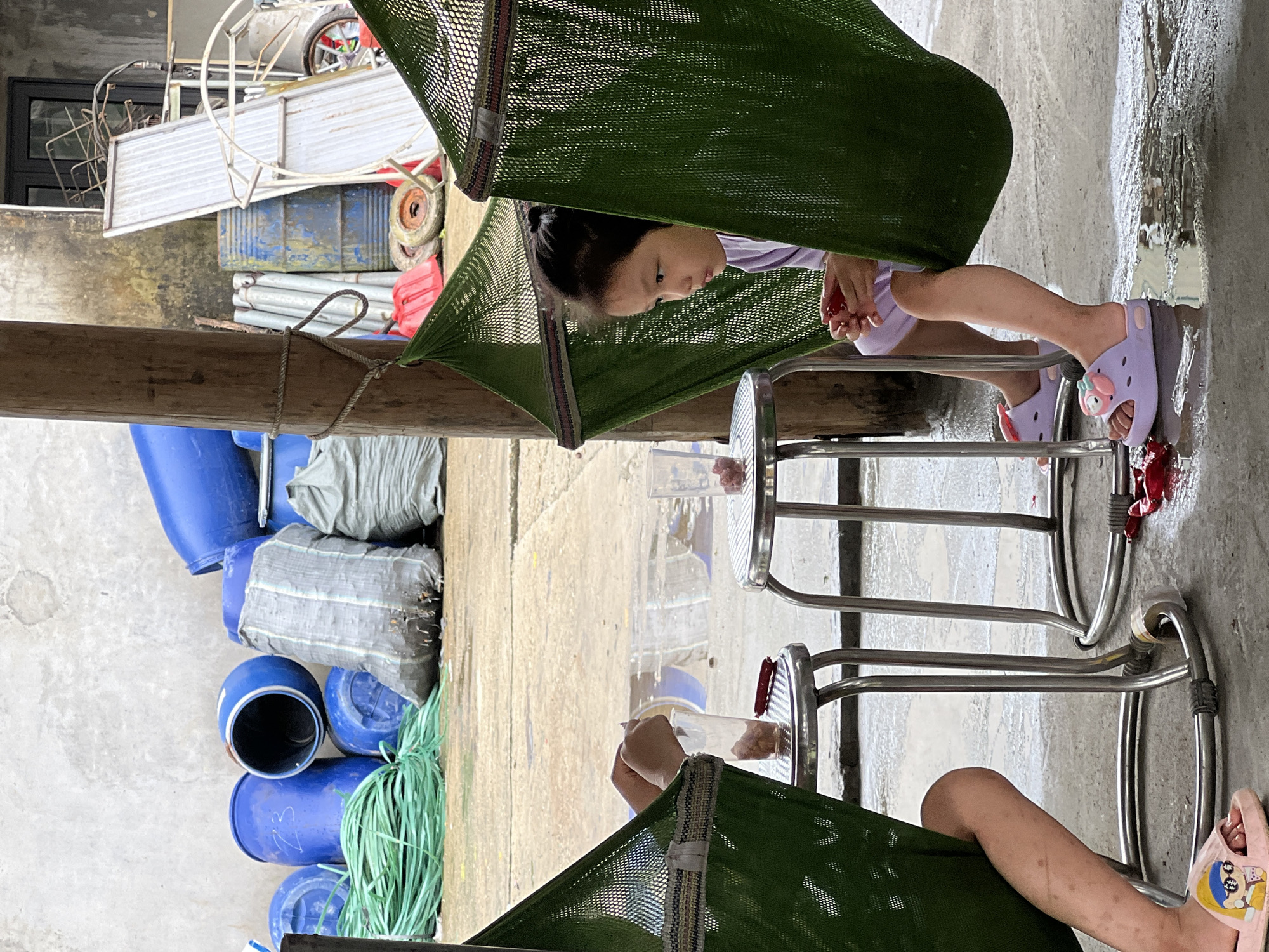 Two children are in hammocks outdoors in Thái Sơn Commune, Vietnam. The child on the right is a young girl with dark hair, wearing a light purple shirt. She is holding a piece of red fruit and looking to her right. The child on the left is only partially visible, with their legs and arms showing. They are also holding something and appear to be interacting with a cup on a small metal table. In the background, there are various objects including blue barrels, green rope, sacks, and metal sheeting, suggesting an industrial or storage area. The ground is wet and reflects the surroundings.